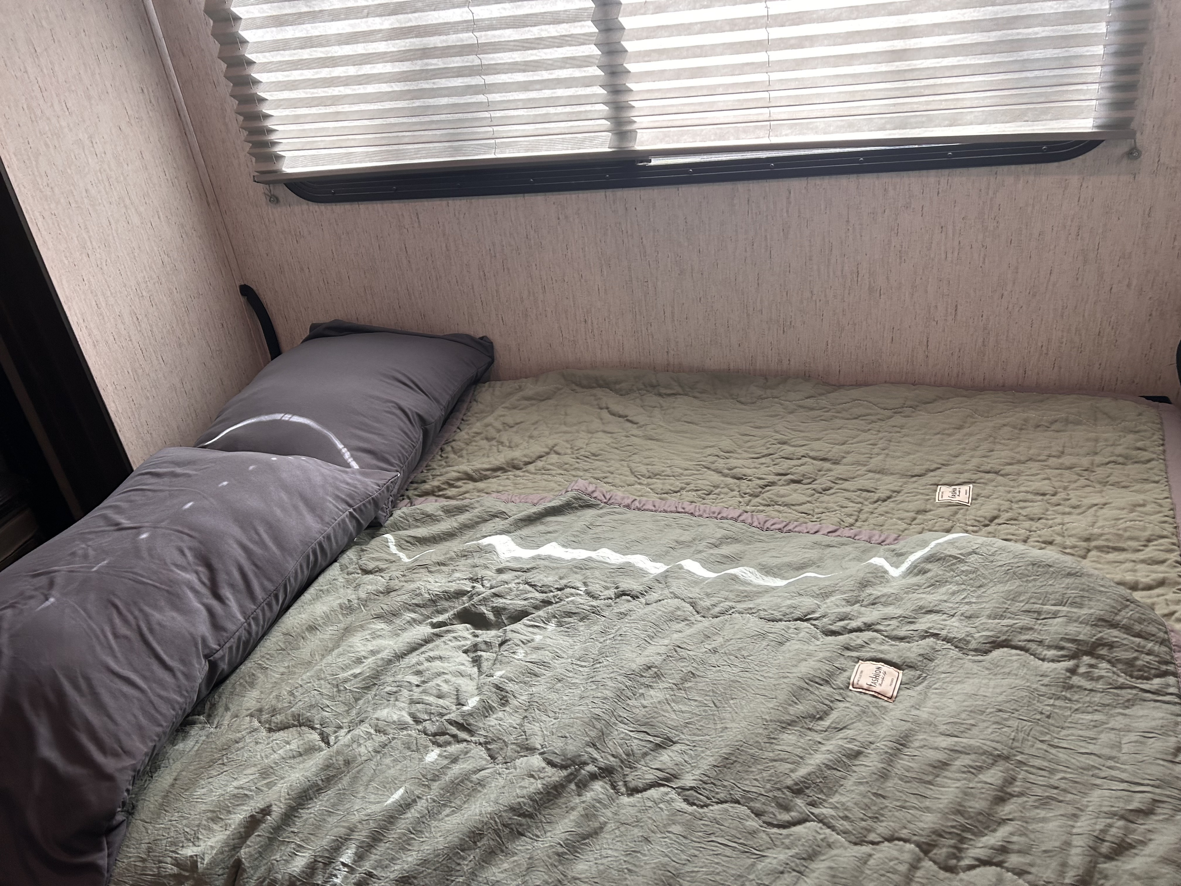 Cozy rv rental bedroom showing a bed with a gray body pillow, green quilted blanket, and pleated window blinds above.. Grand Design Momentum 2022