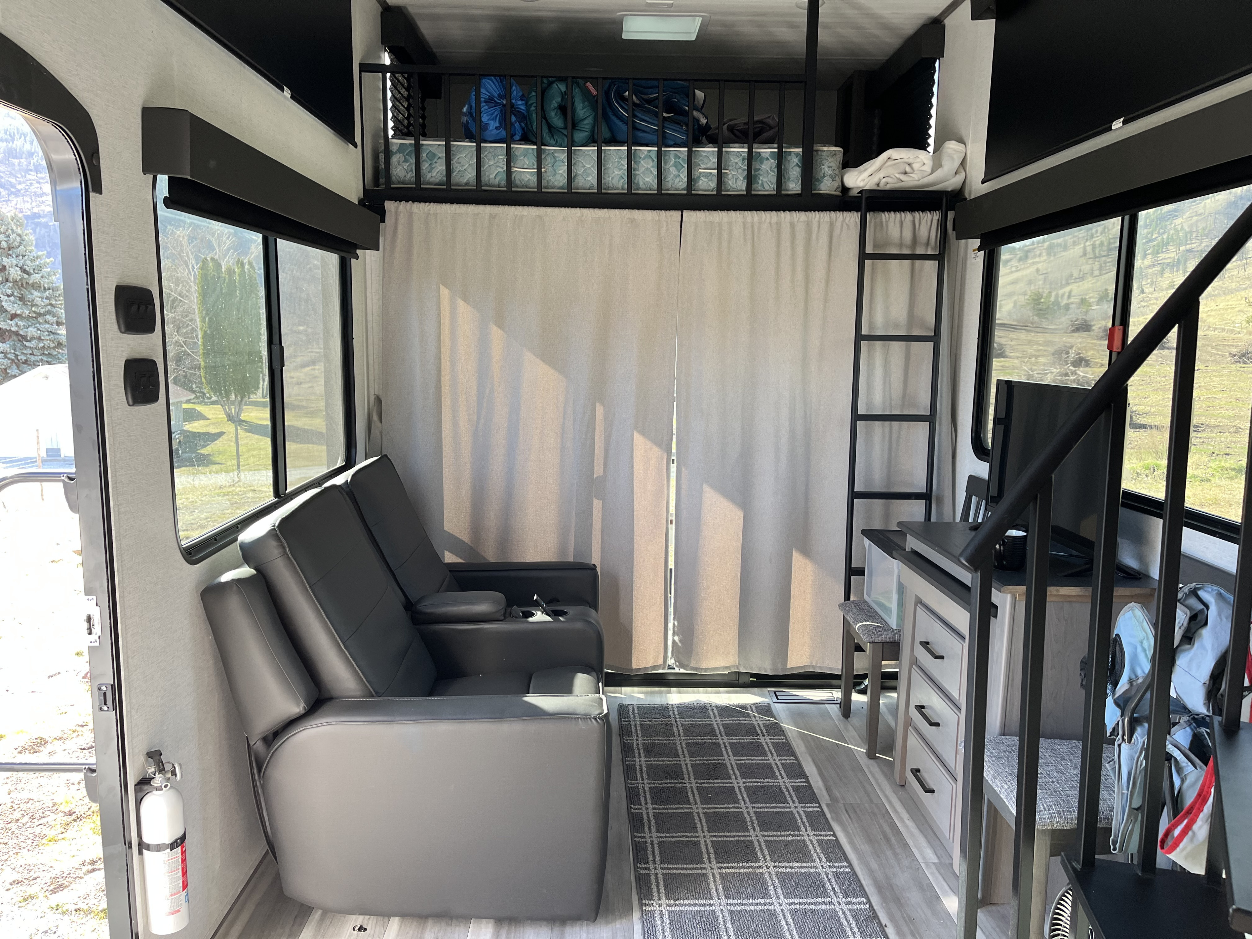 Inside this modern **rv rental**: reclining sofa, loft bunk with ladder, desk/TV, large windows with views, and a fire extinguisher.. Keystone RV Hideout 2024