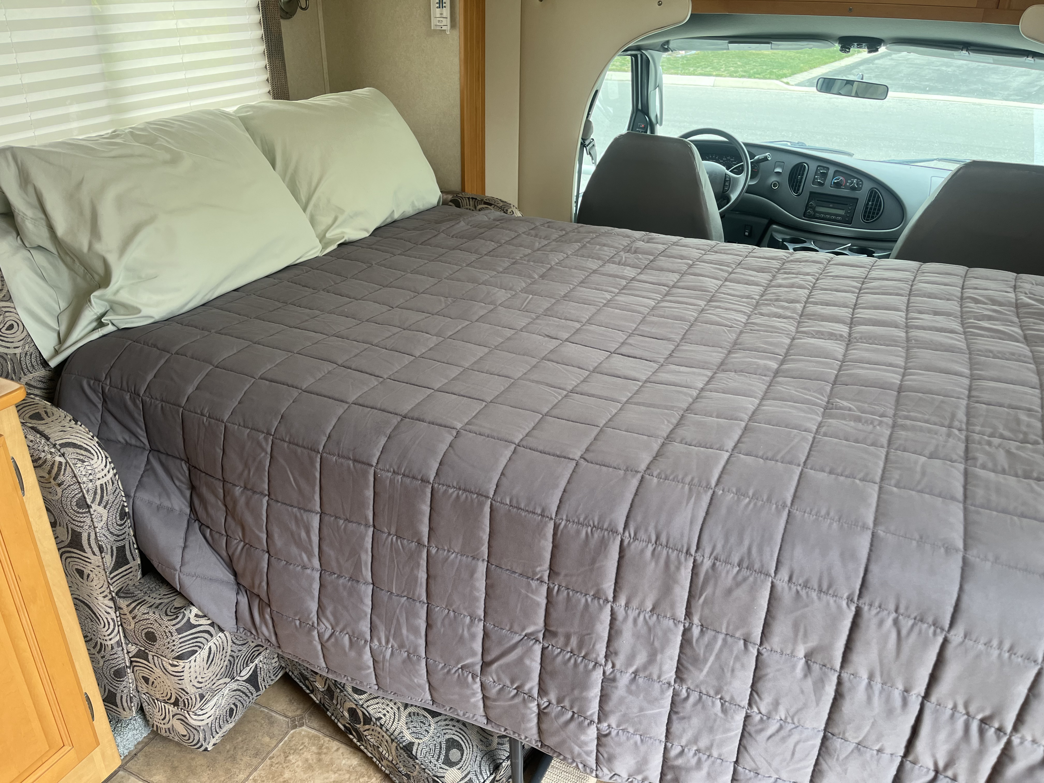 RV rental interior shows a cozy cab-over bunk bed with gray quilt and green pillows, extending over the driver's cockpit.. Triple E Regency 2006
