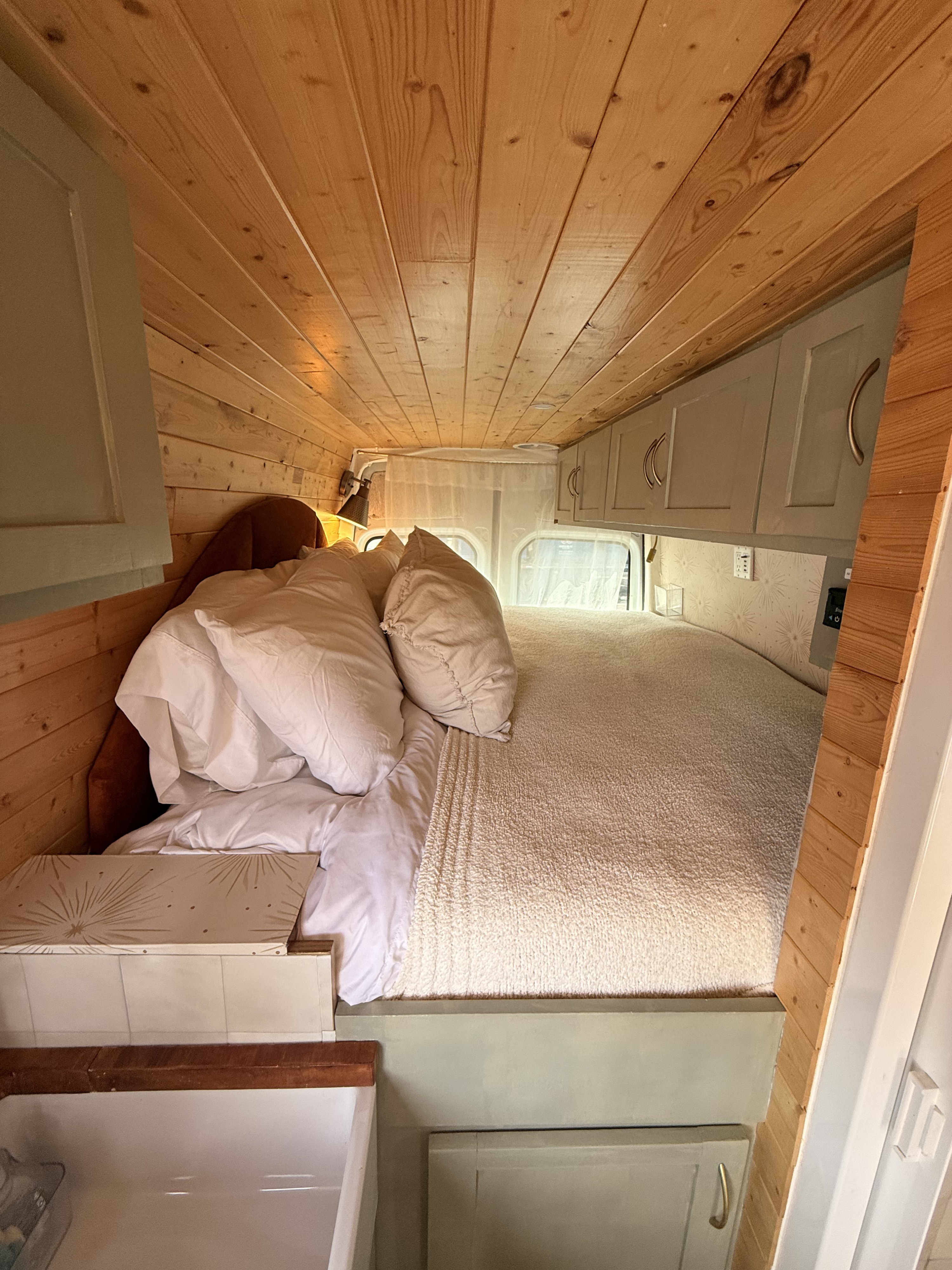 Cozy RV rental bedroom with rustic wood paneling, soft bed, overhead storage, and a small sink.. Mercedes-Benz Sprinter 2011