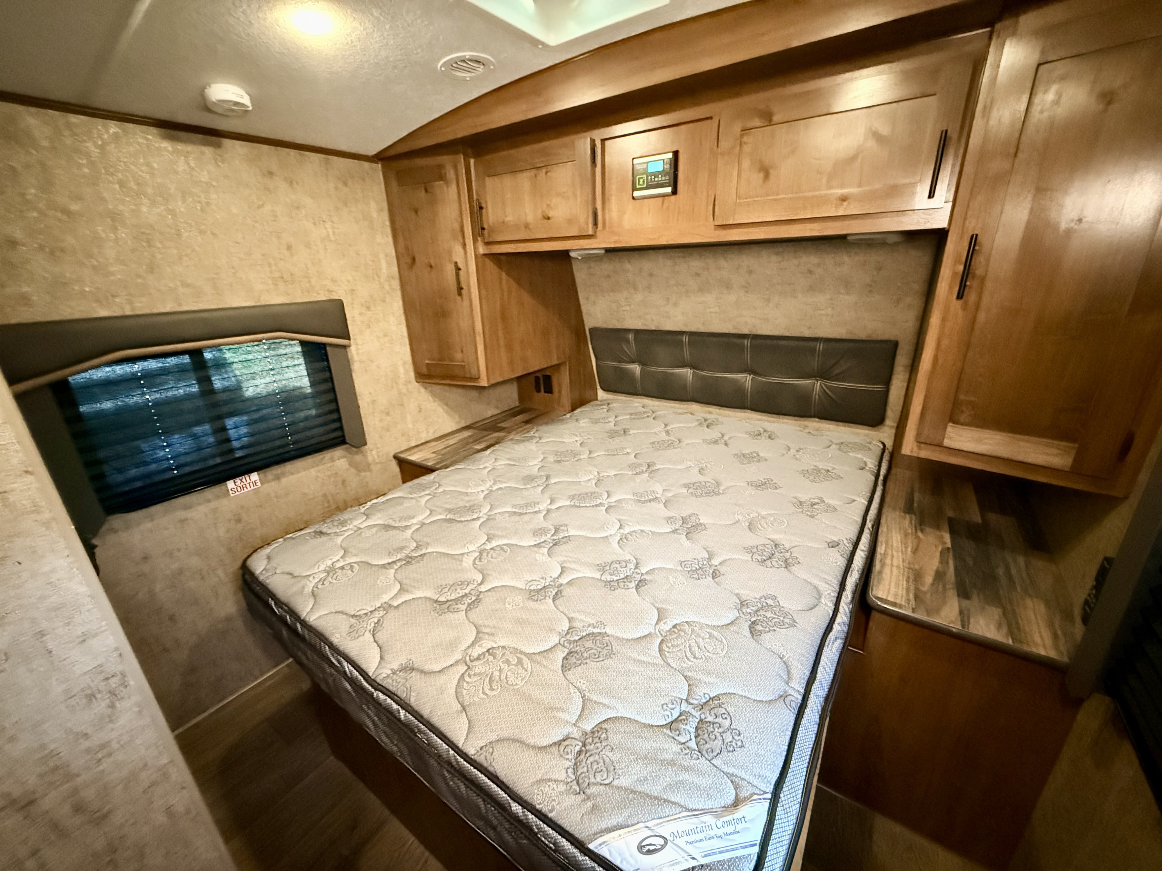 Interior of an RV rental bedroom with a large bed, brown headboard, wooden overhead cabinets, and a window with blinds.. Outdoors RV Mountain 2021