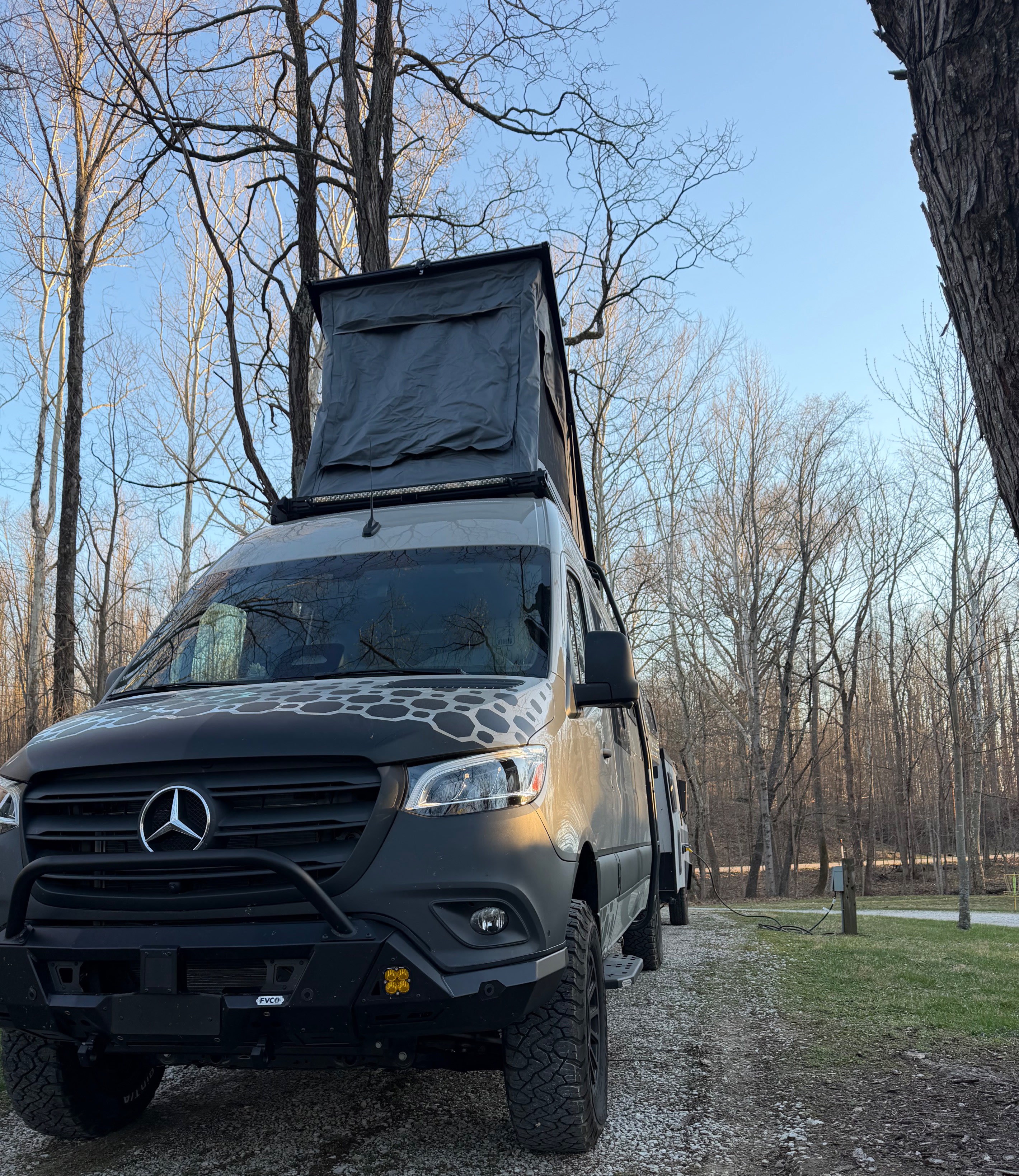 Adventure-ready Mercedes Sprinter rv rental with pop-top tent, parked on a gravel path in a wooded campsite.. Mercedes-Benz Sprinter 2026