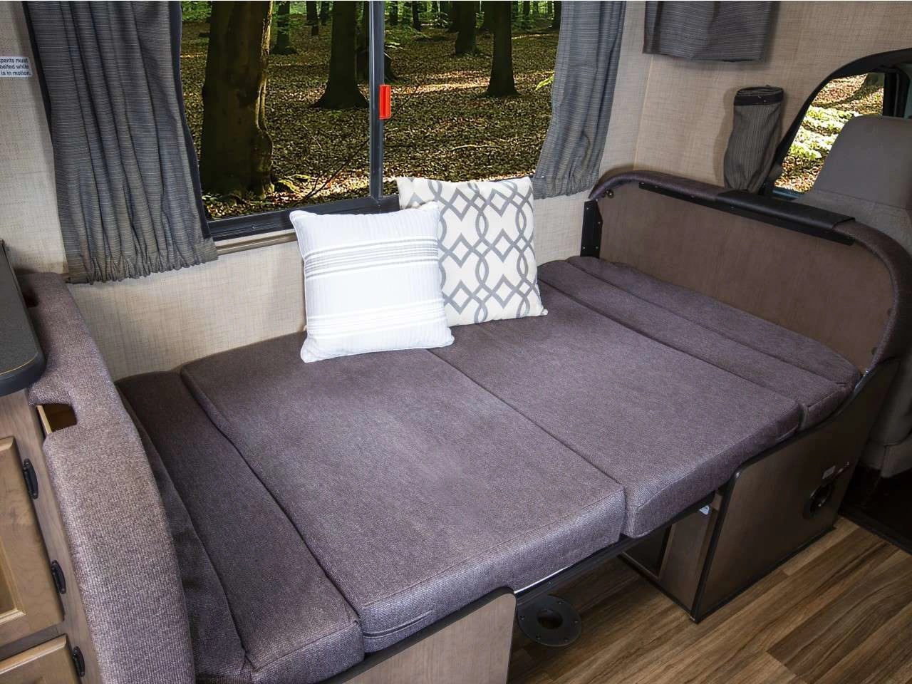 Cozy RV rental interior: A gray sofa bed with decorative pillows and a forest view from the window.. Thor Majestic 2019