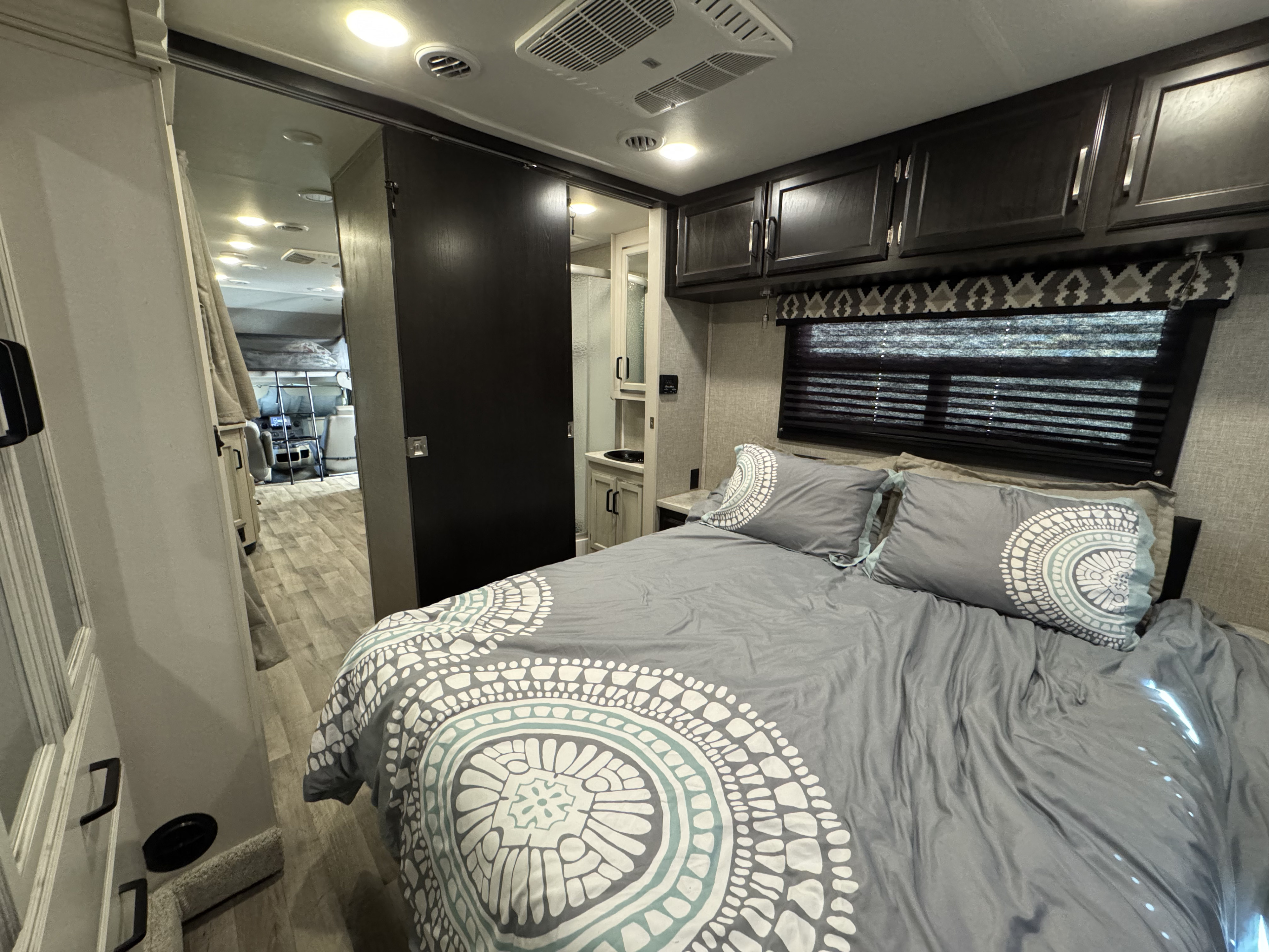 Spacious RV rental bedroom with patterned gray bedding, an adjacent bathroom, and a glimpse into the living area with bunk beds.. Jayco Redhawk 2022