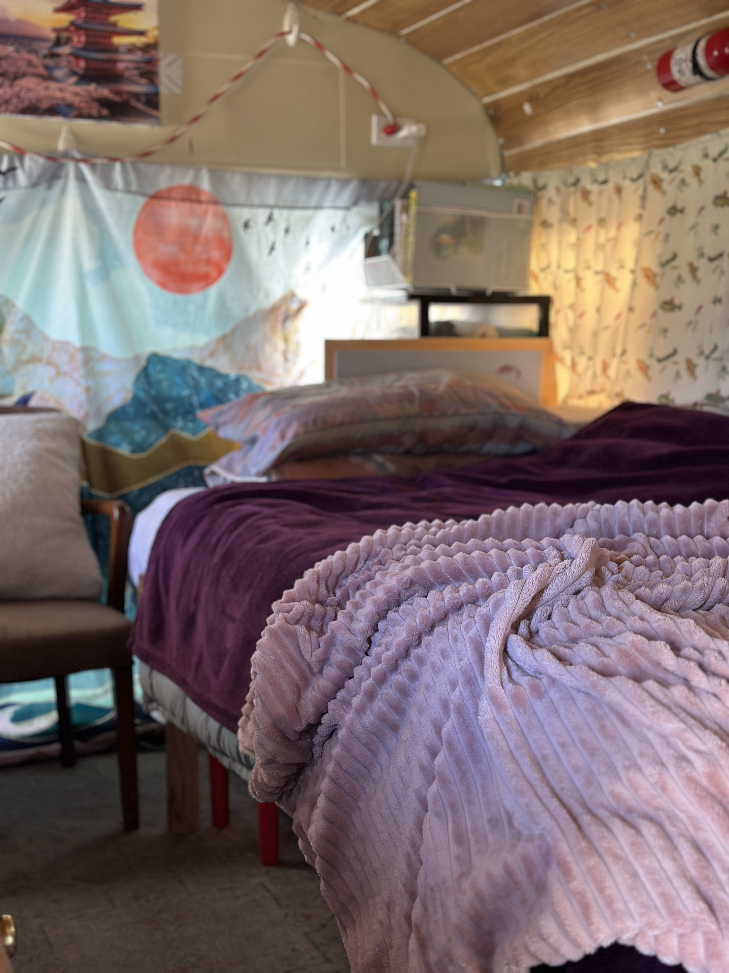 Inside a cozy RV rental: a plush bed with purple and pink blankets, nature tapestry, and wood-paneled ceiling.. GMC Vandura 1986
