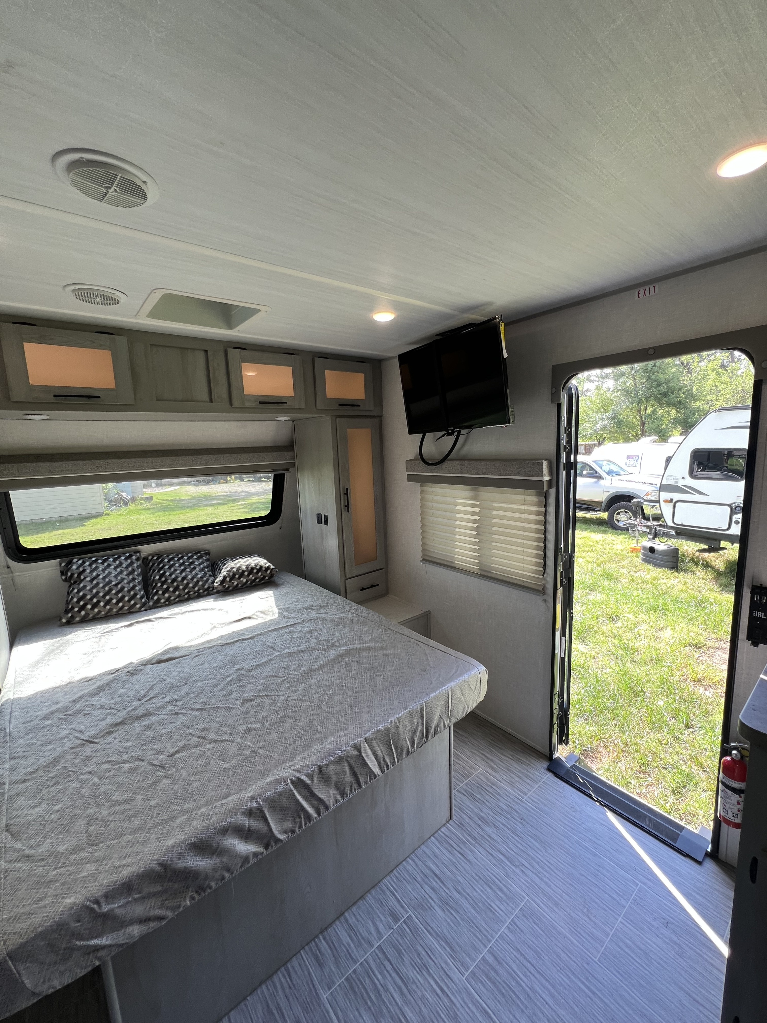 RV rental bedroom with a bed, patterned pillows, wall-mounted TV, and an open door to a sunny campsite.. Forest River Ibex 2021