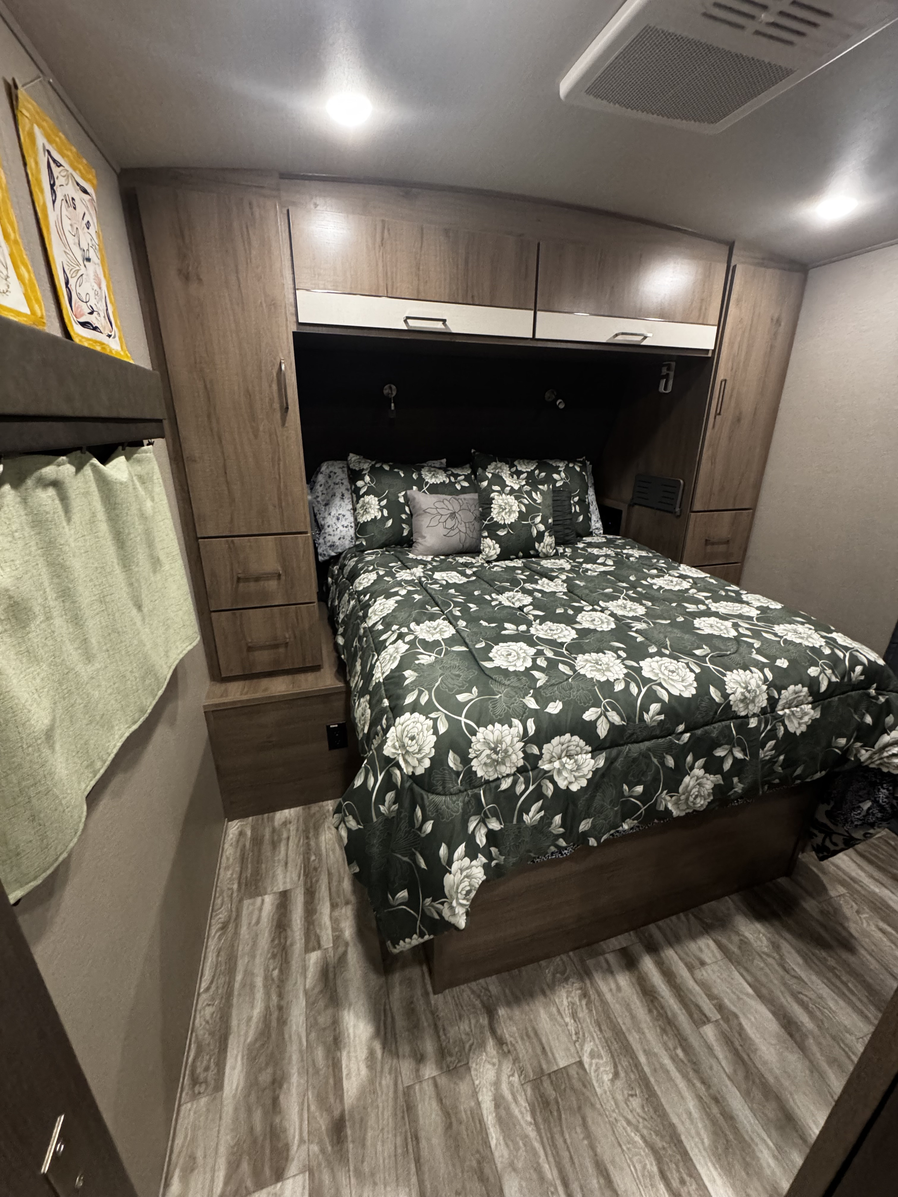 Cozy RV rental bedroom features a dark green floral comforter, wood-toned cabinetry, built-in lights, and wood-look flooring.. Grand Design Imagine 2022