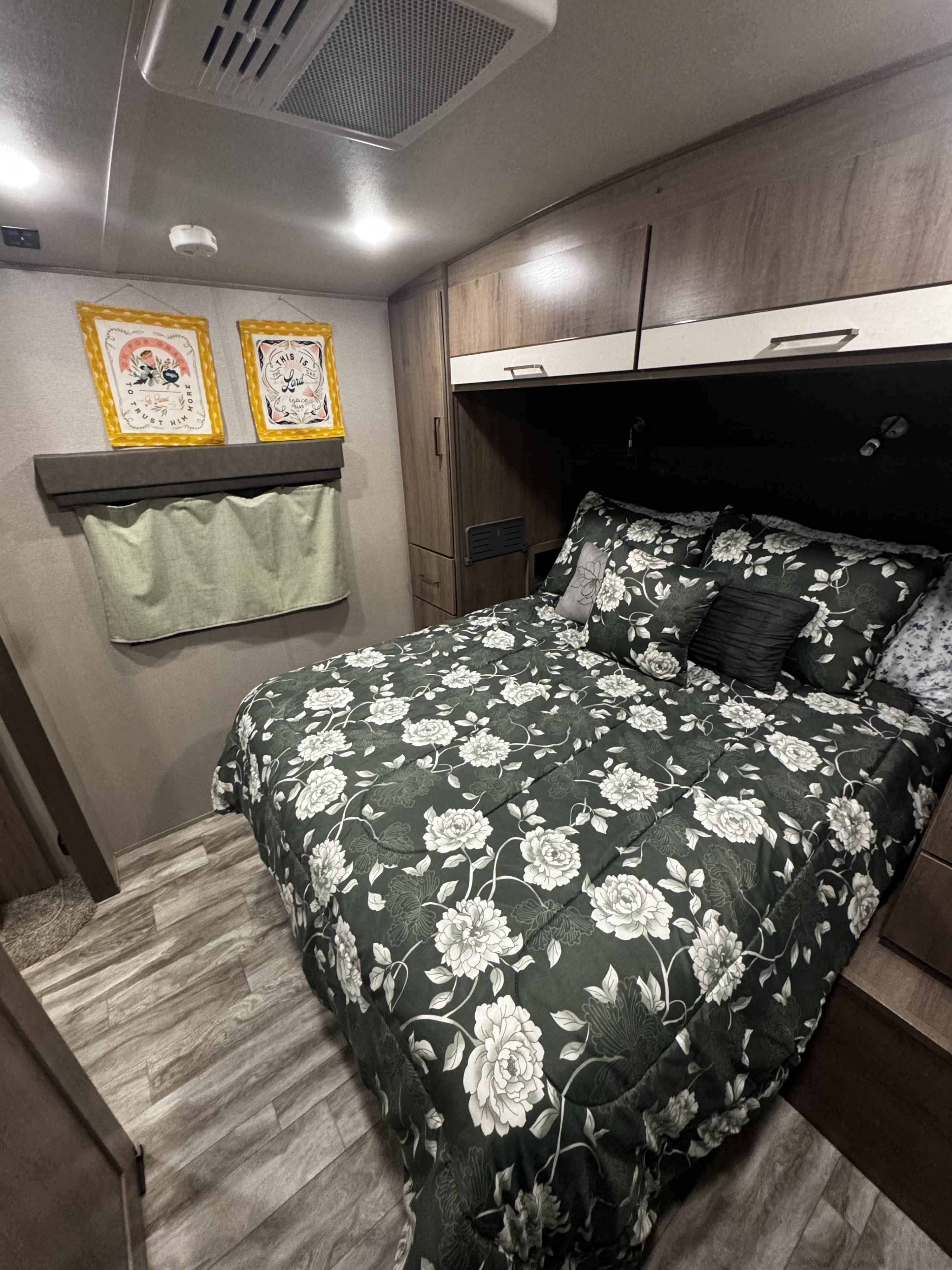 This inviting RV rental bedroom features a dark green floral comforter, overhead storage, and warm lighting, creating a cozy retreat.. Grand Design Imagine 2022