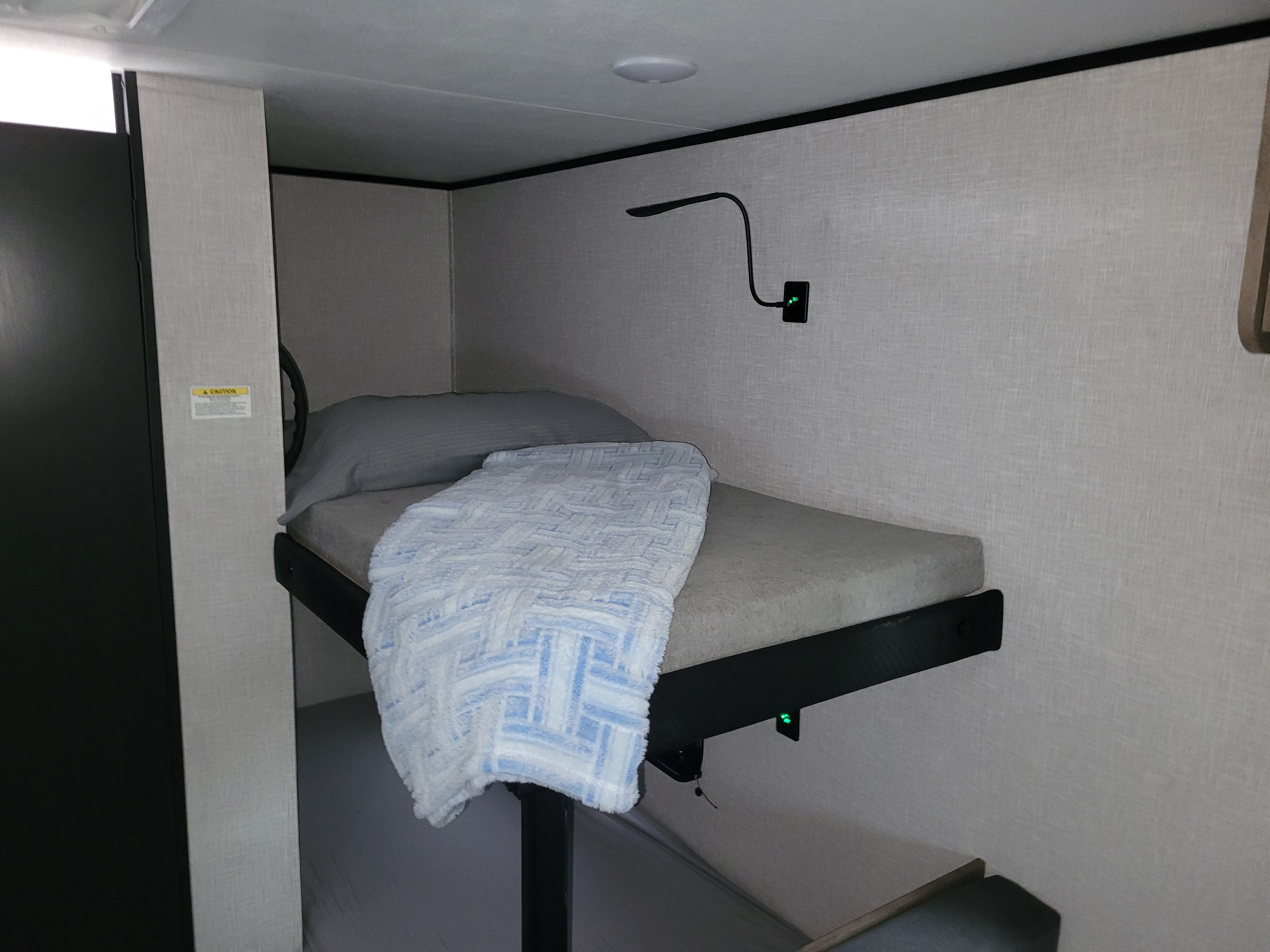 RV rental bunk bed: a grey pillow, blue/white blanket, and wall-mounted reading light on textured walls. Cozy, compact sleeping space.. Jayco Jay Flight SLX 2025