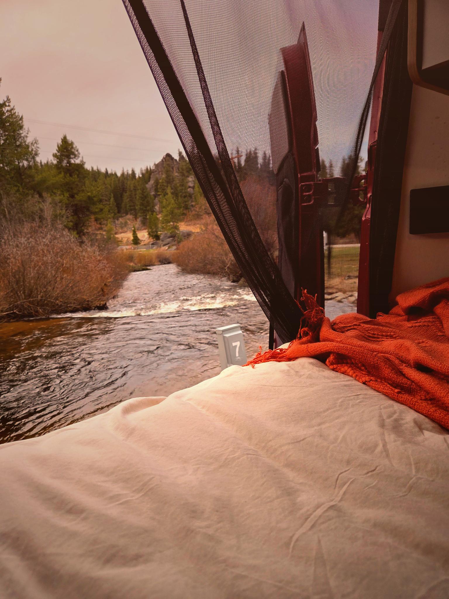 Cozy RV rental bed with an orange blanket and bug net, looking out at a flowing river, pine forest, and rock formations.. Winnebago Solis 2022
