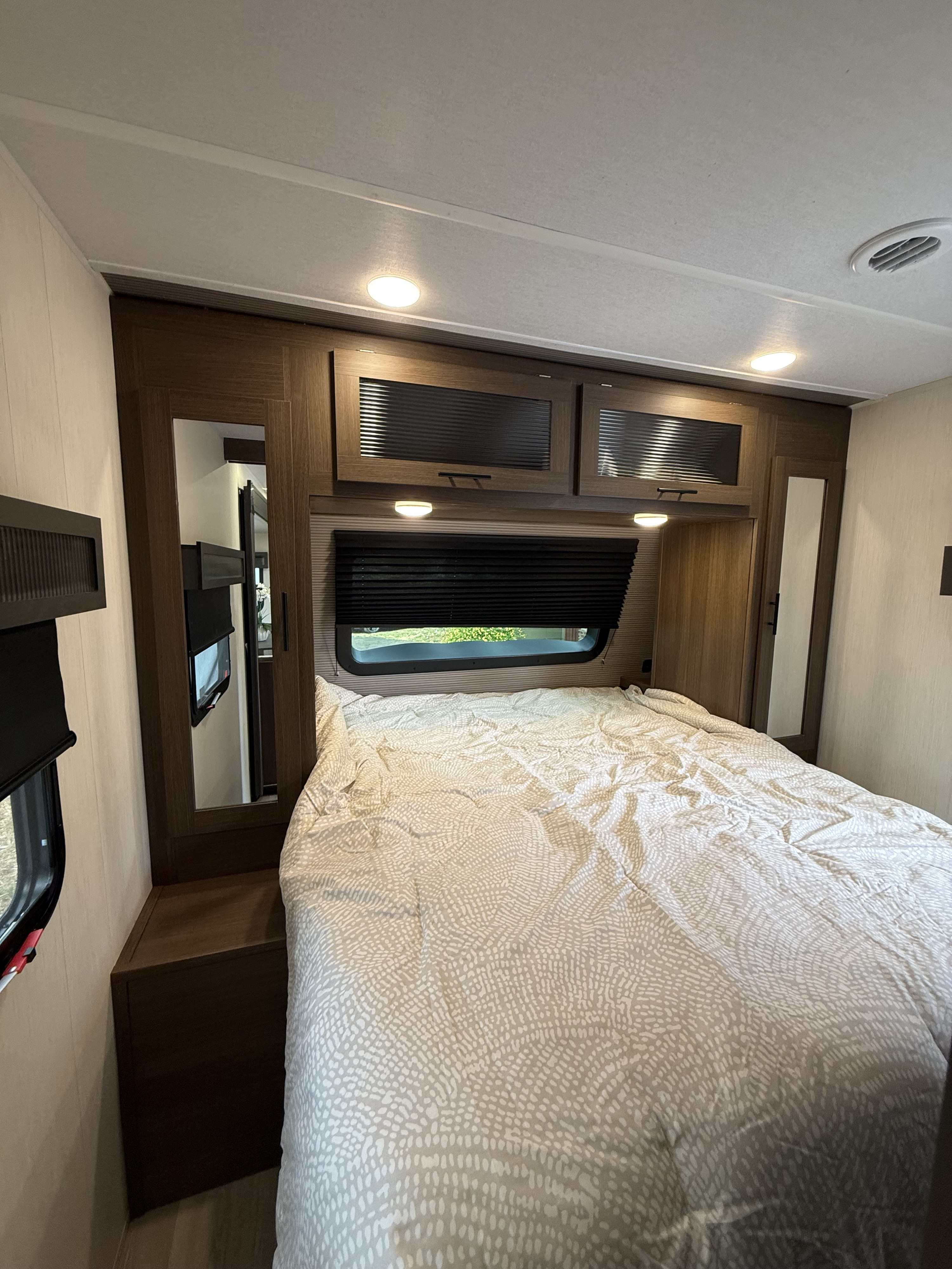 RV rental bedroom featuring a large bed with patterned duvet, dark wood cabinets, window with blind, and ceiling lights.. Forest River Wildcat One 241DBEDS 2026