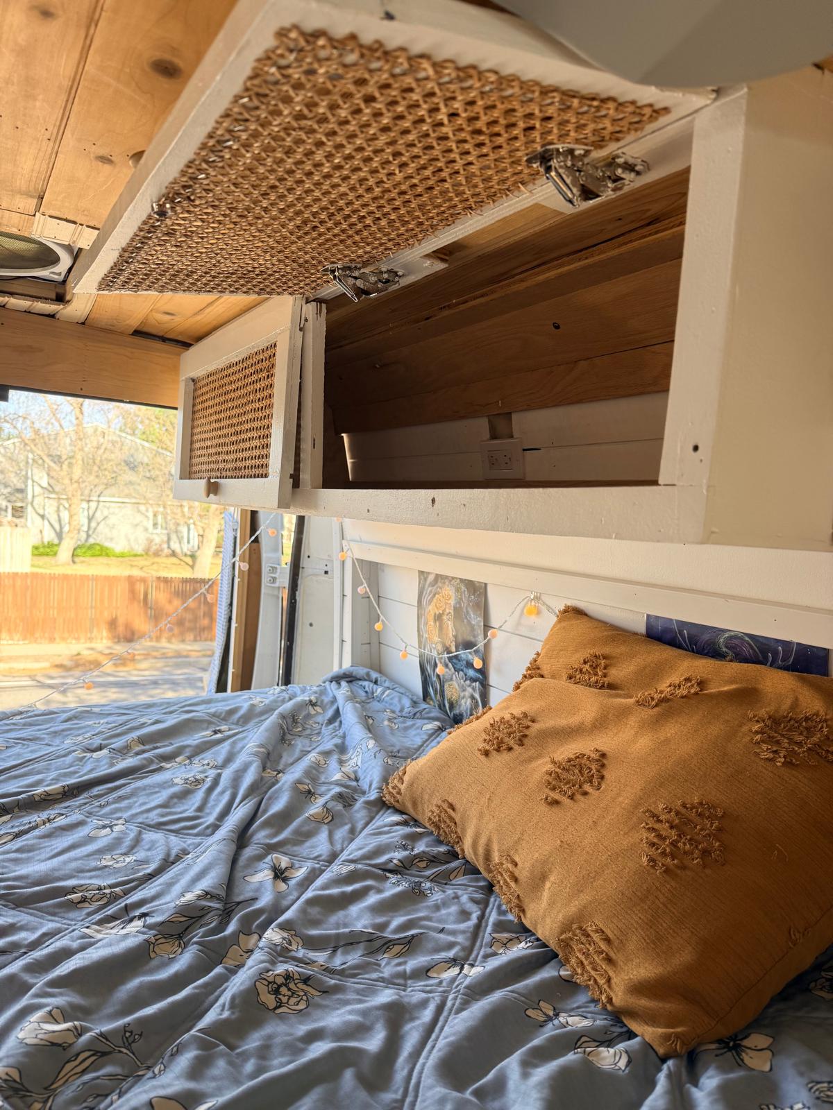 Cozy camper van interior featuring custom overhead cabinets with rattan inserts, a bed with a floral duvet, and fairy lights. Ideal for an RV rental.. Ram Promaster 2019