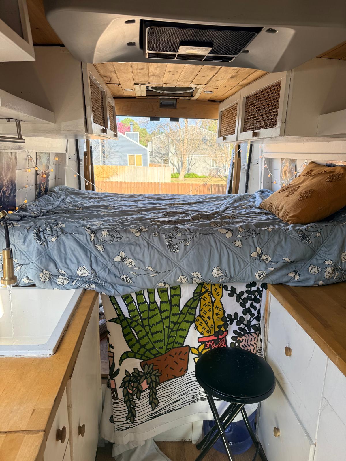 Cozy rv rental interior with bed, flowing faucet, and nature-themed decor looking out to a suburban backyard.. Ram Promaster 2019