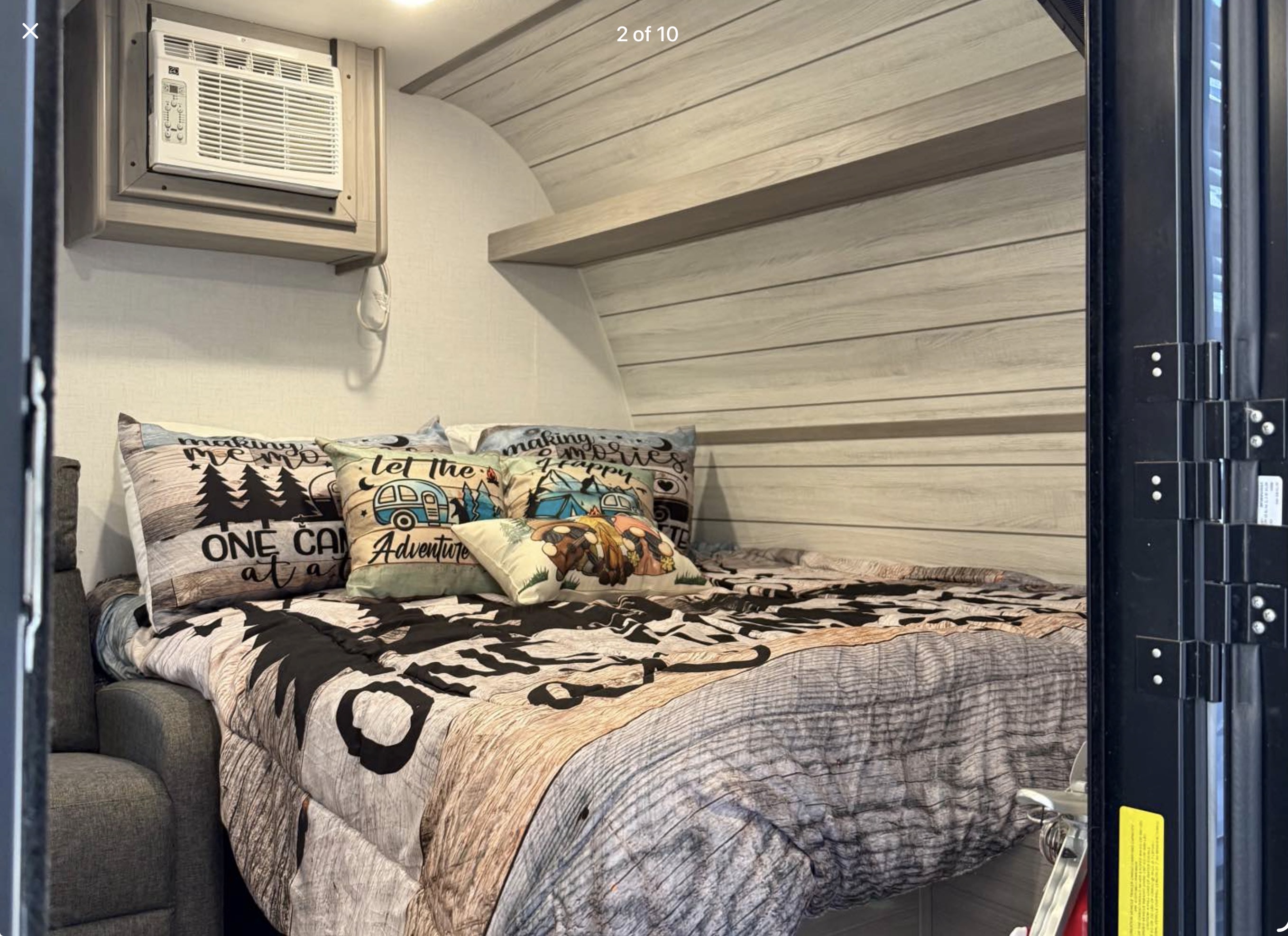 Interior of an rv rental features a cozy bed with adventure-themed bedding, a wall-mounted AC unit, and wood-slat walls.. Dutchmen Coleman Lantern LT 2024
