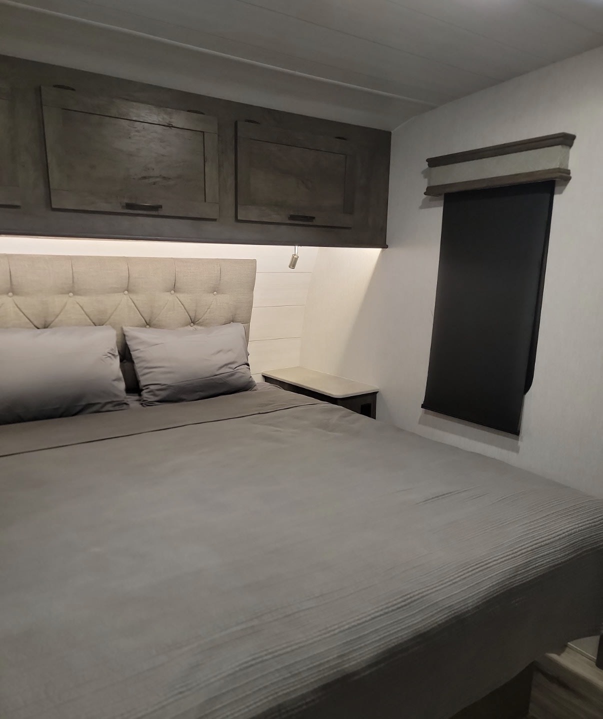 A modern, compact bedroom setup in an RV rental, with a gray bed, tufted headboard, and overhead cabinets.. Forest River Sandstorm 2022