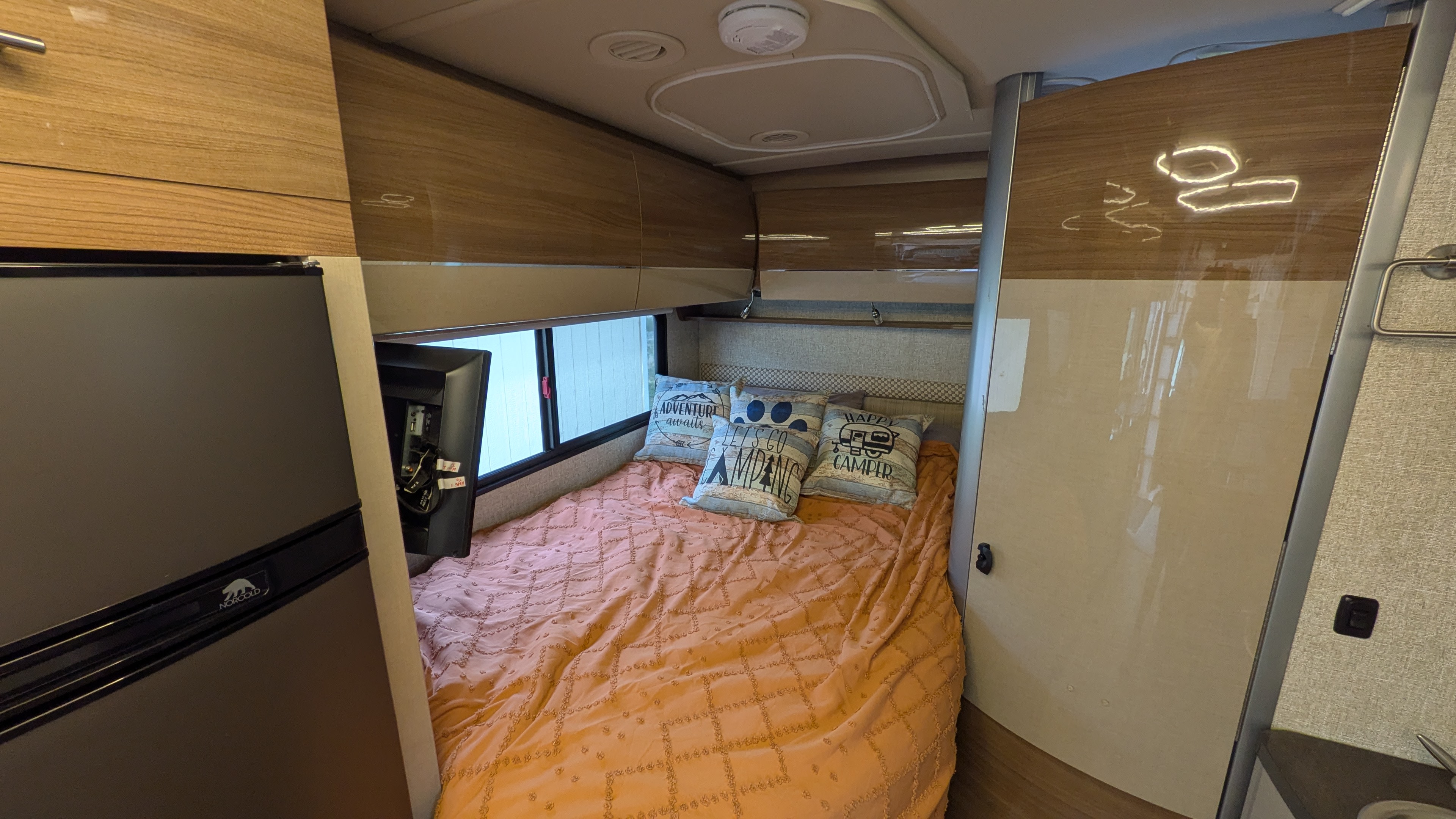 Cozy RV rental bedroom featuring an orange bed with decorative pillows, a window, overhead cabinets, and a refrigerator.. Winnebago View 2017
