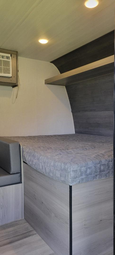 Modern RV rental interior: cozy sleeping area with patterned bed, grey wood-look cabinetry, AC unit, and bright overhead lights.. Keystone RV Hideout 2024