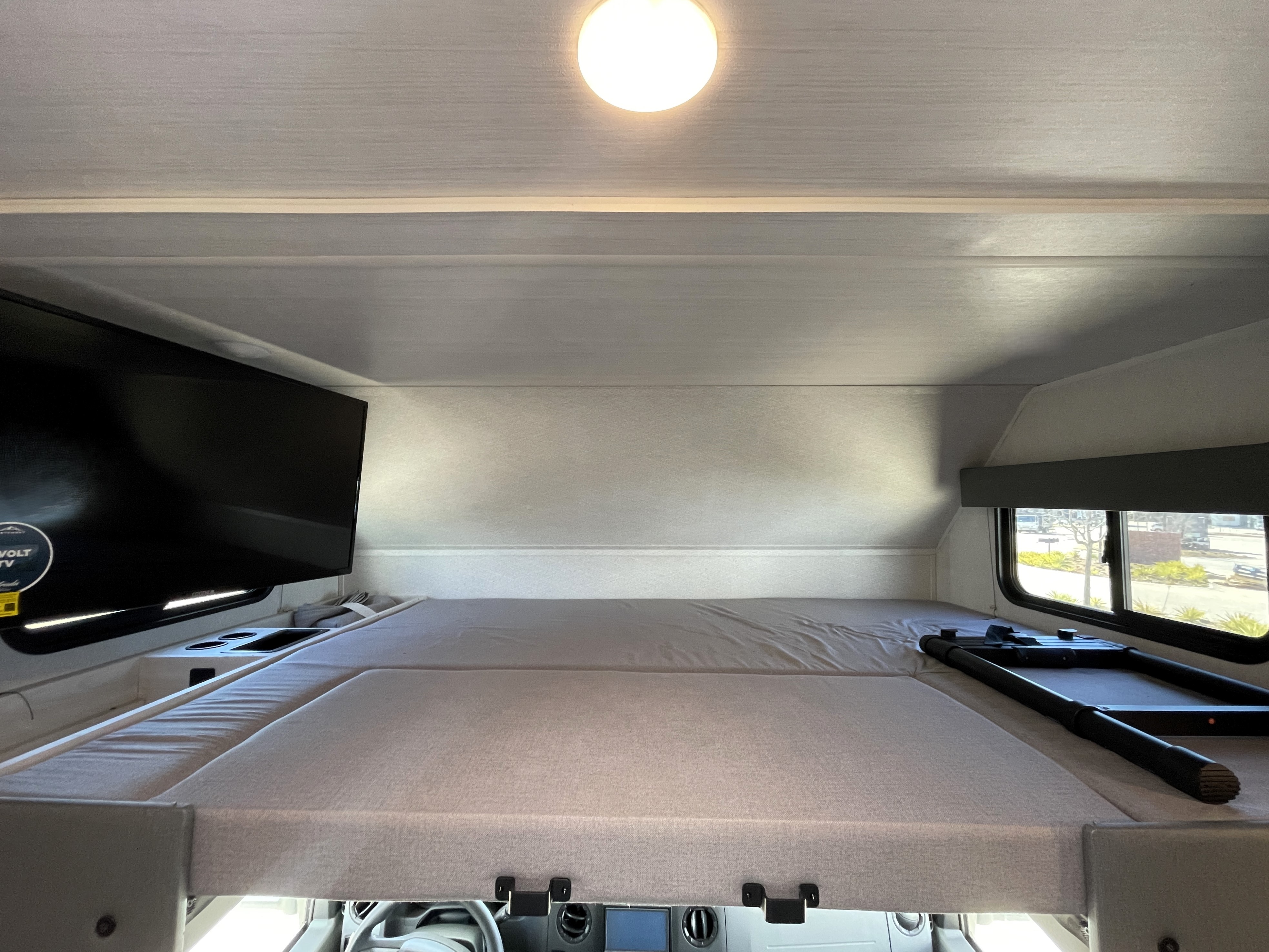 Overhead bunk bed in an RV rental. Features a TV on the left, a window on the right, ceiling light, and an entry ladder.. East to West Entrada 2024