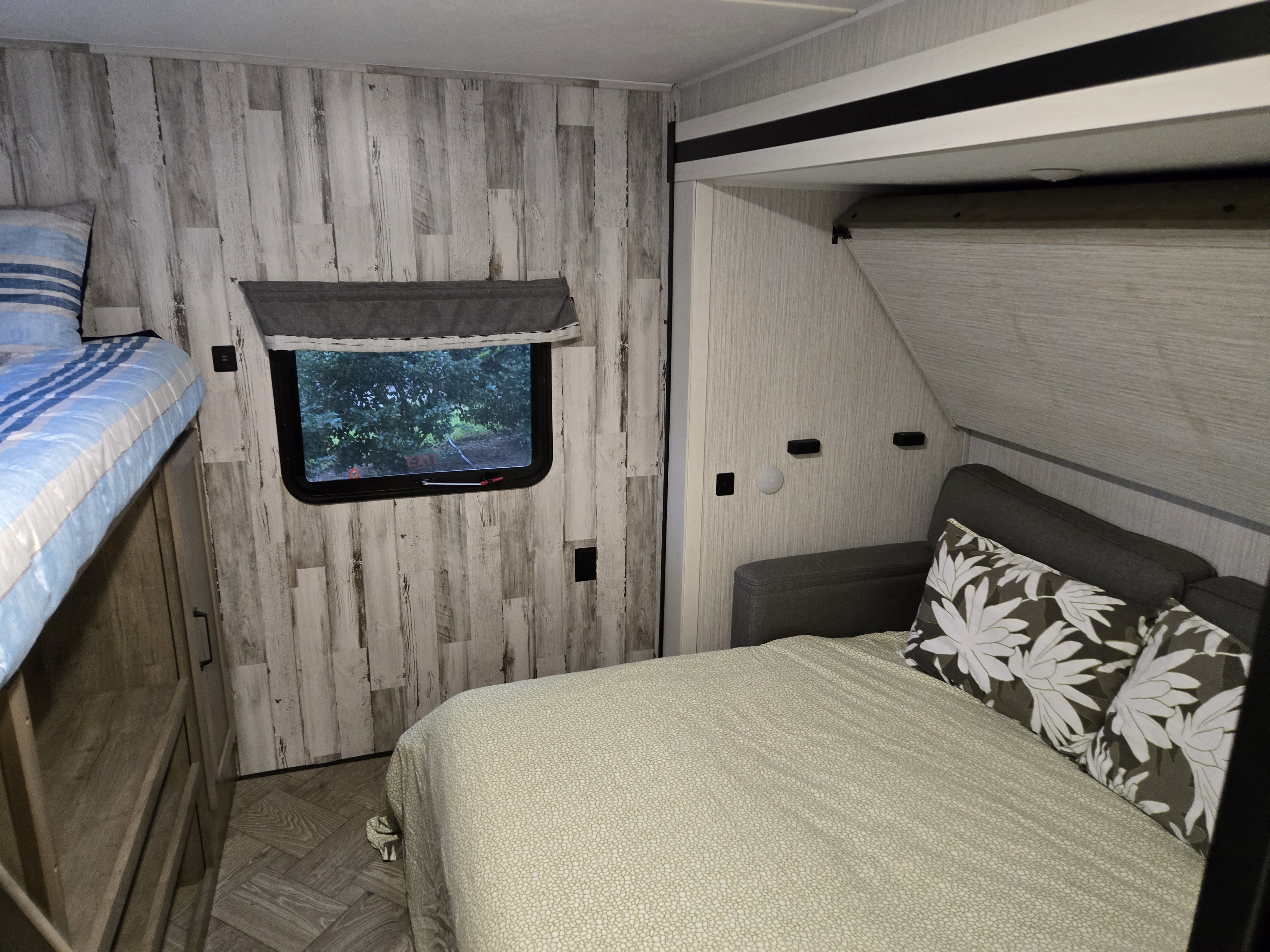 Cozy RV rental bedroom with a comfy lower bed, floral pillows, and an upper bunk bed. Features rustic wood-look walls and a scenic window.. Forest River Palomino Puma 2022