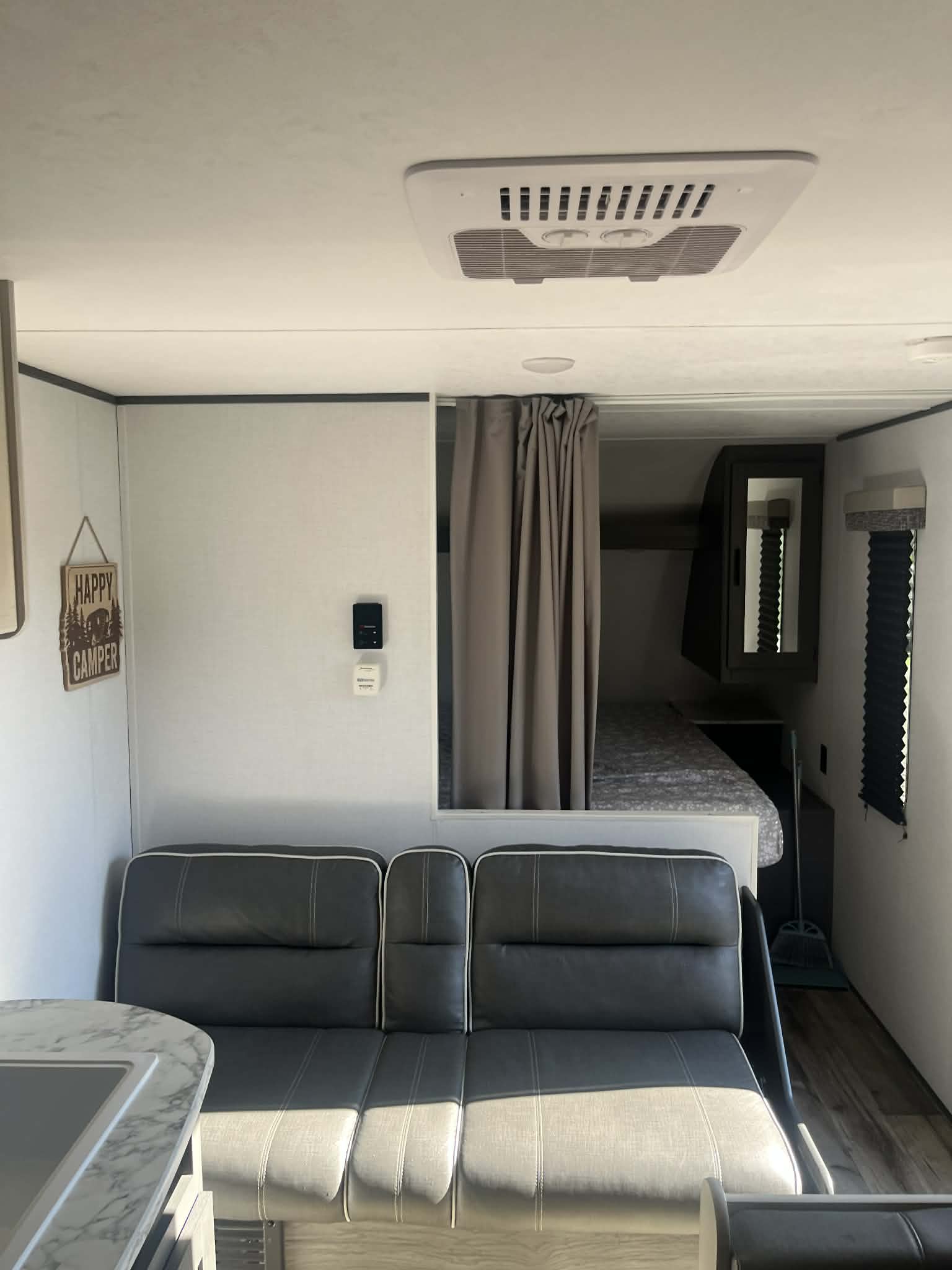 RV rental interior featuring a gray sofa/dinette, AC unit, "Happy Camper" sign, and a curtained bedroom area.. Dutchmen Aspen Trail 2023