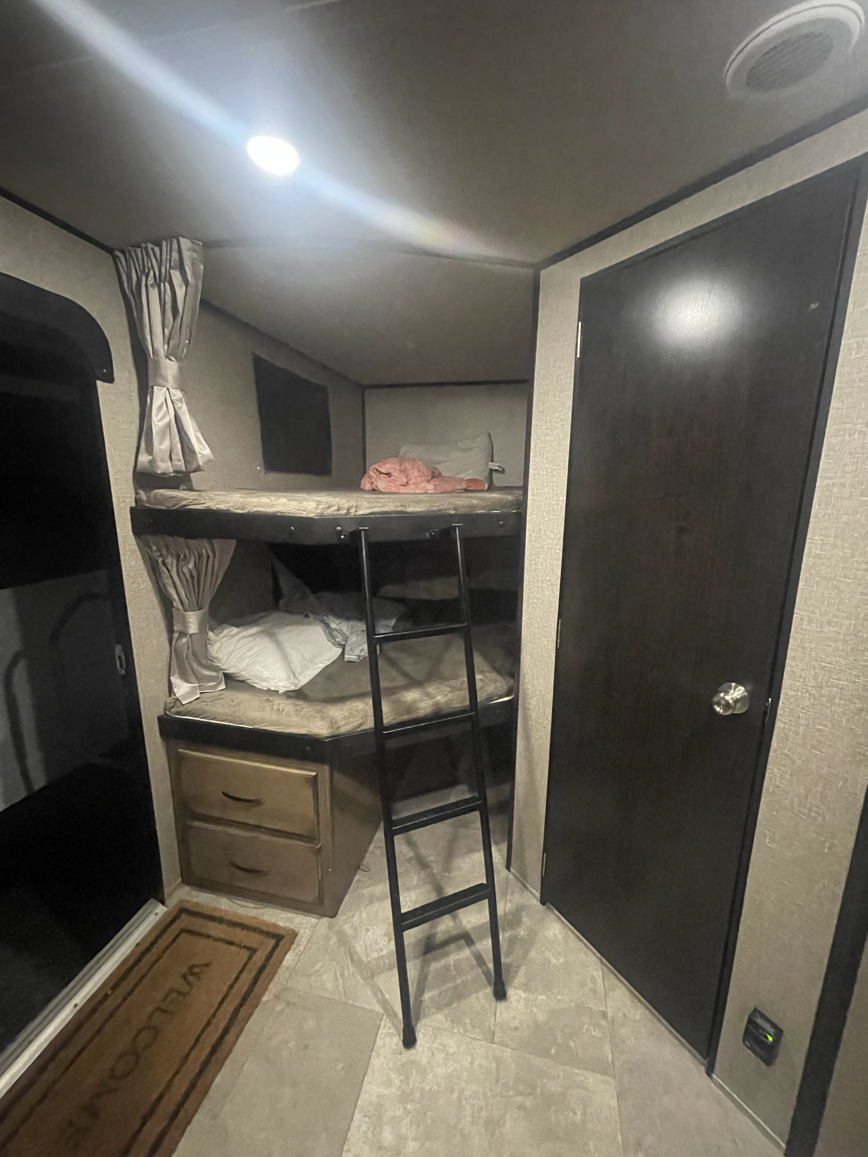 Inside an RV rental: bunk beds with a ladder, side curtains, storage, and a "welcome" mat at the entry.. Grand Design Transcend 2019