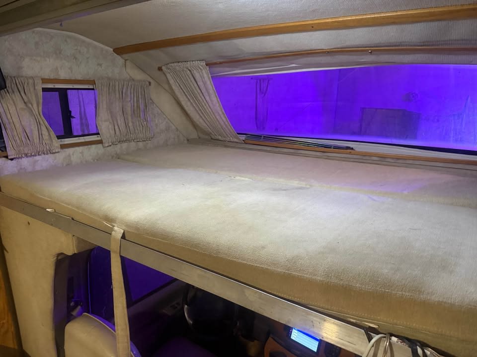 Over-cab bunk sleeping area in an RV rental, showing mattress, curtains, windows, and purple exterior light.. Gulf Stream Gulfstream 1999