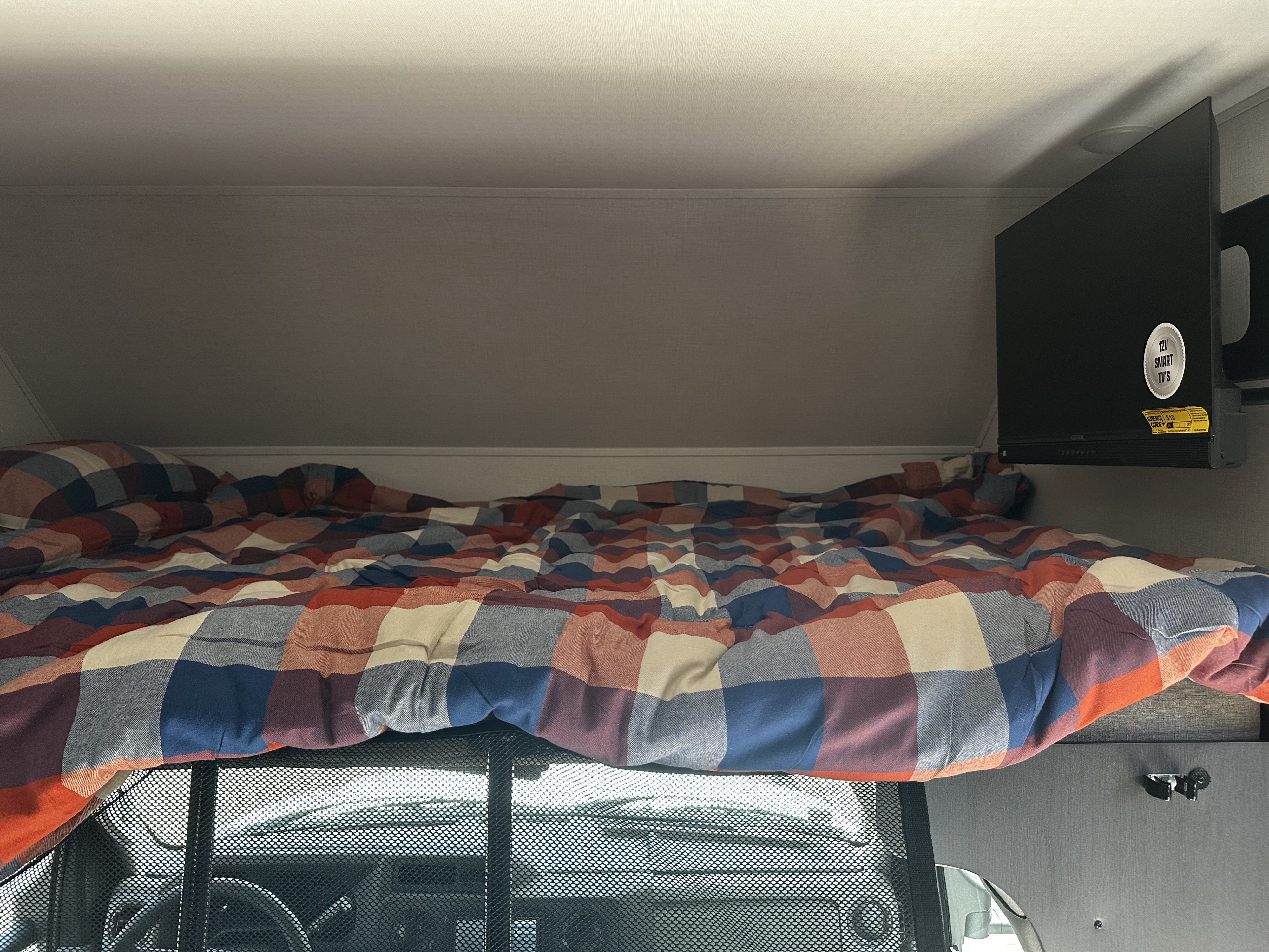 View of an RV rental's cozy overhead sleeping area, featuring a plaid bed, safety net, and mounted TV.. Forester LE 2026