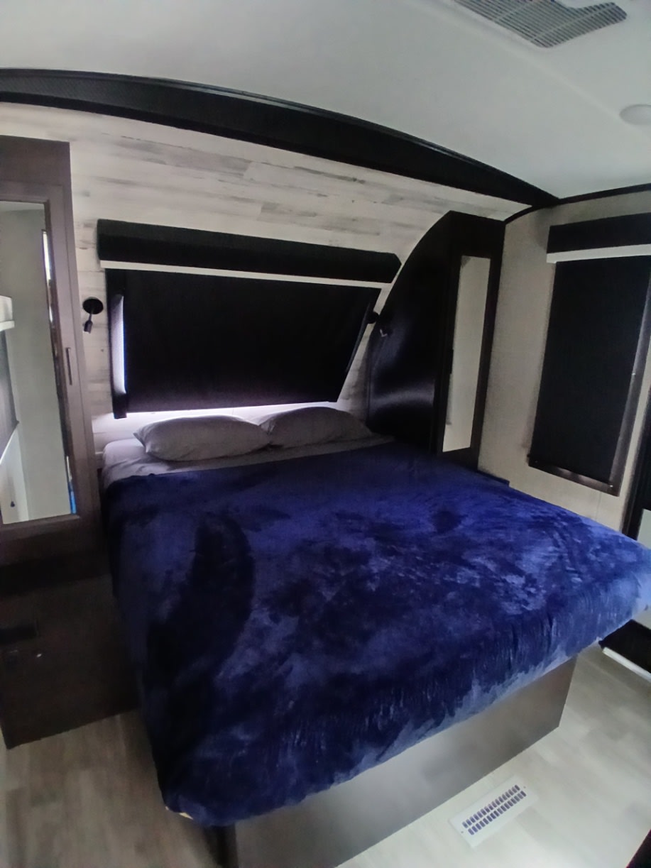 Cozy RV rental bedroom with a blue blanket, blackout blinds, and modern light wood-paneled walls. Perfect for your trip.. Jayco White Hawk 2023