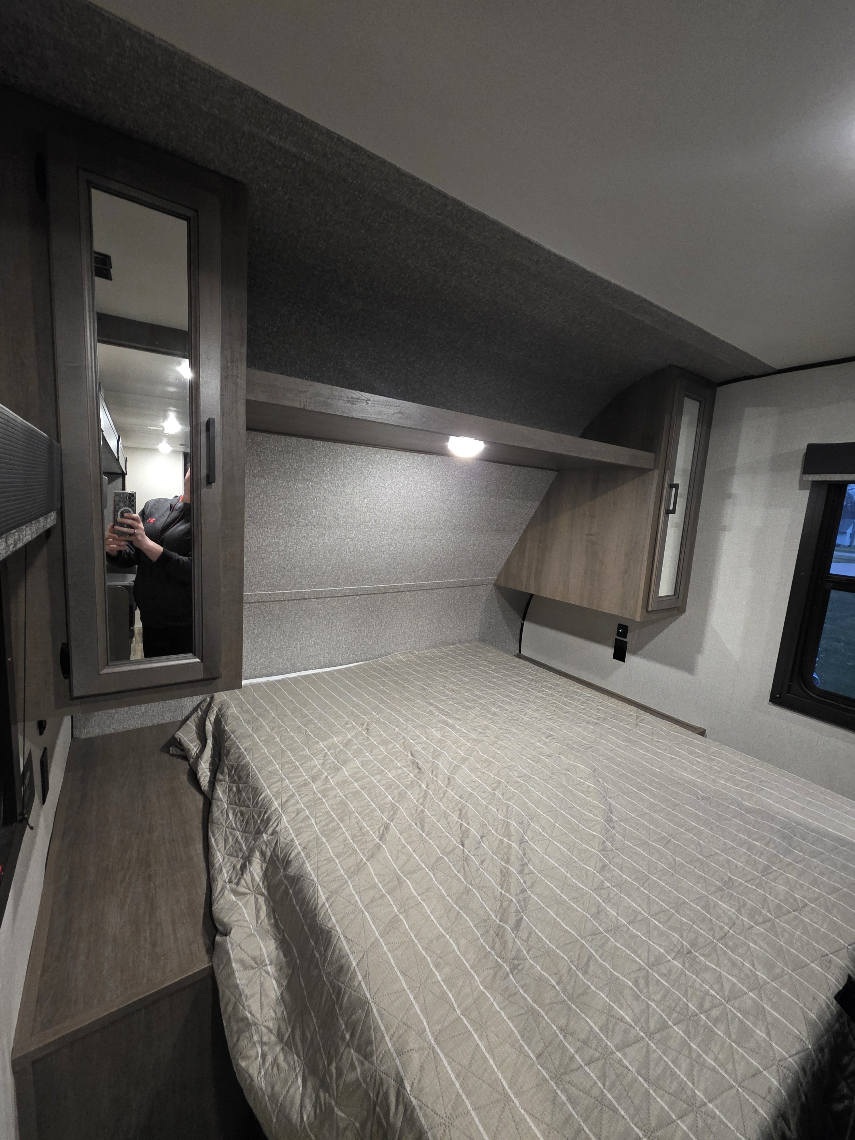 Inside this rv rental bedroom, a quilted bed is flanked by mirrored cabinets, with an overhead shelf/light and a window.. Starcraft Autumn Ridge 2022