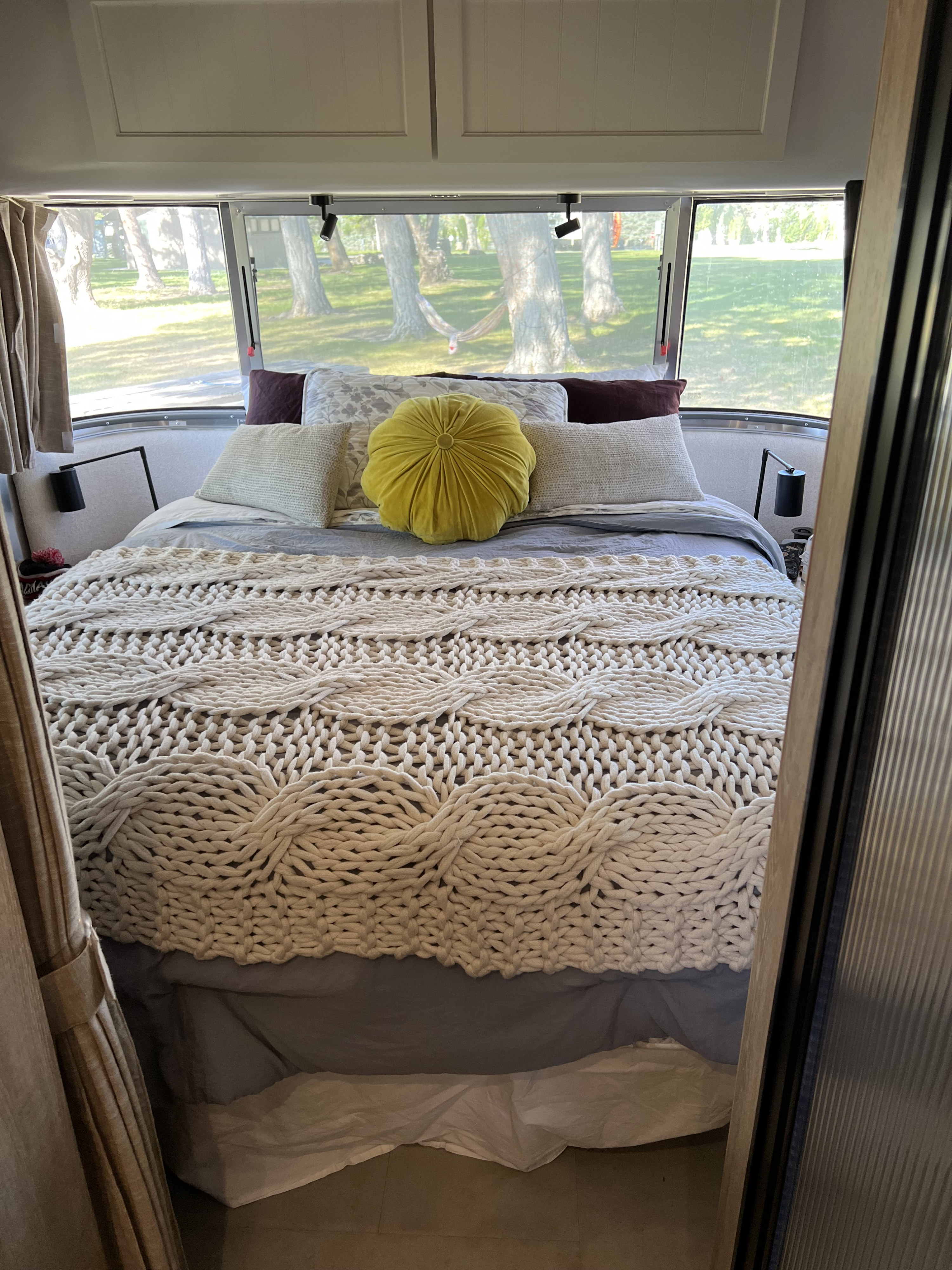 Cozy RV rental bedroom featuring a bed with a chunky knit blanket & yellow pillow, offering serene views through its windows.. Airstream Pottery Barn 2023