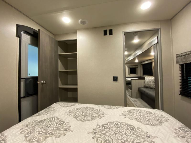 RV rental bedroom: patterned bed, open door, shelves, and a mirror reflecting the living area with seating. Compact design.. Coachmen catalina 2022