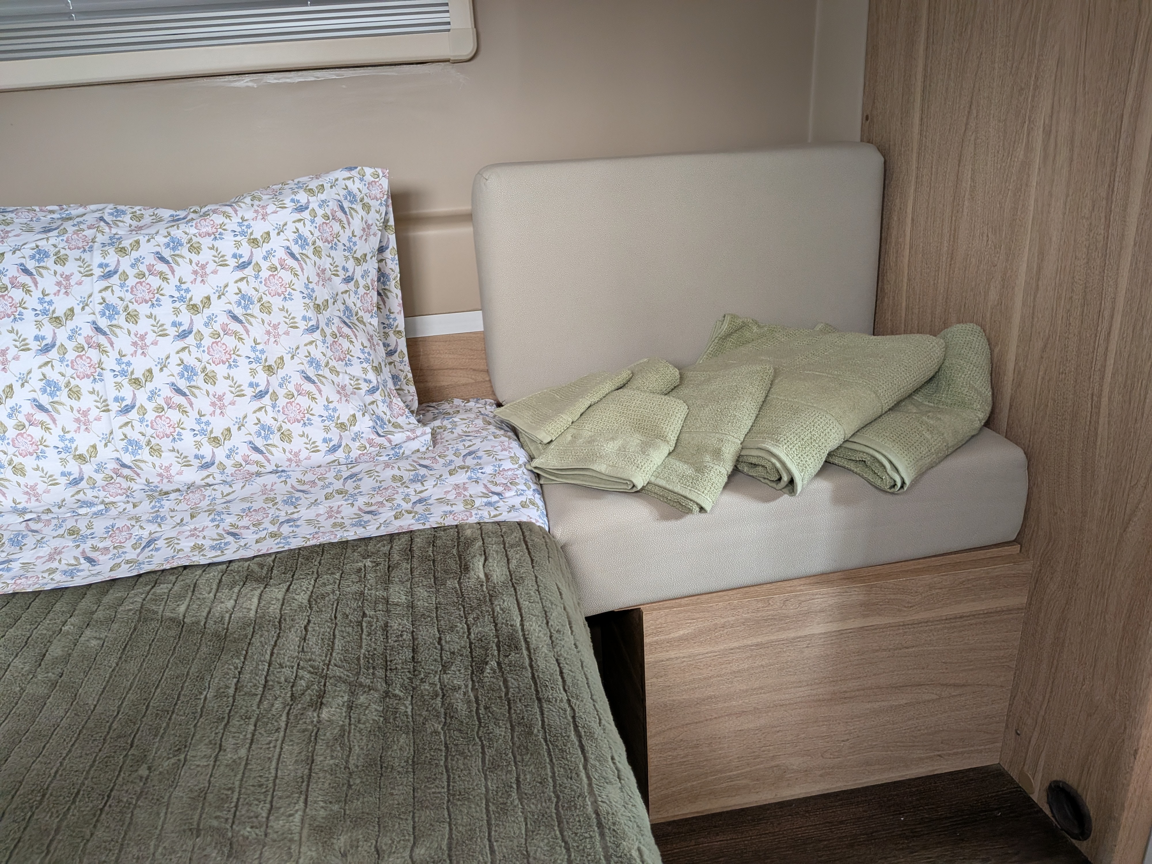 Compact RV rental bedroom with floral bedding, green blanket, and folded towels on a beige cushioned seat.. Hymer Hymer Class B 2018