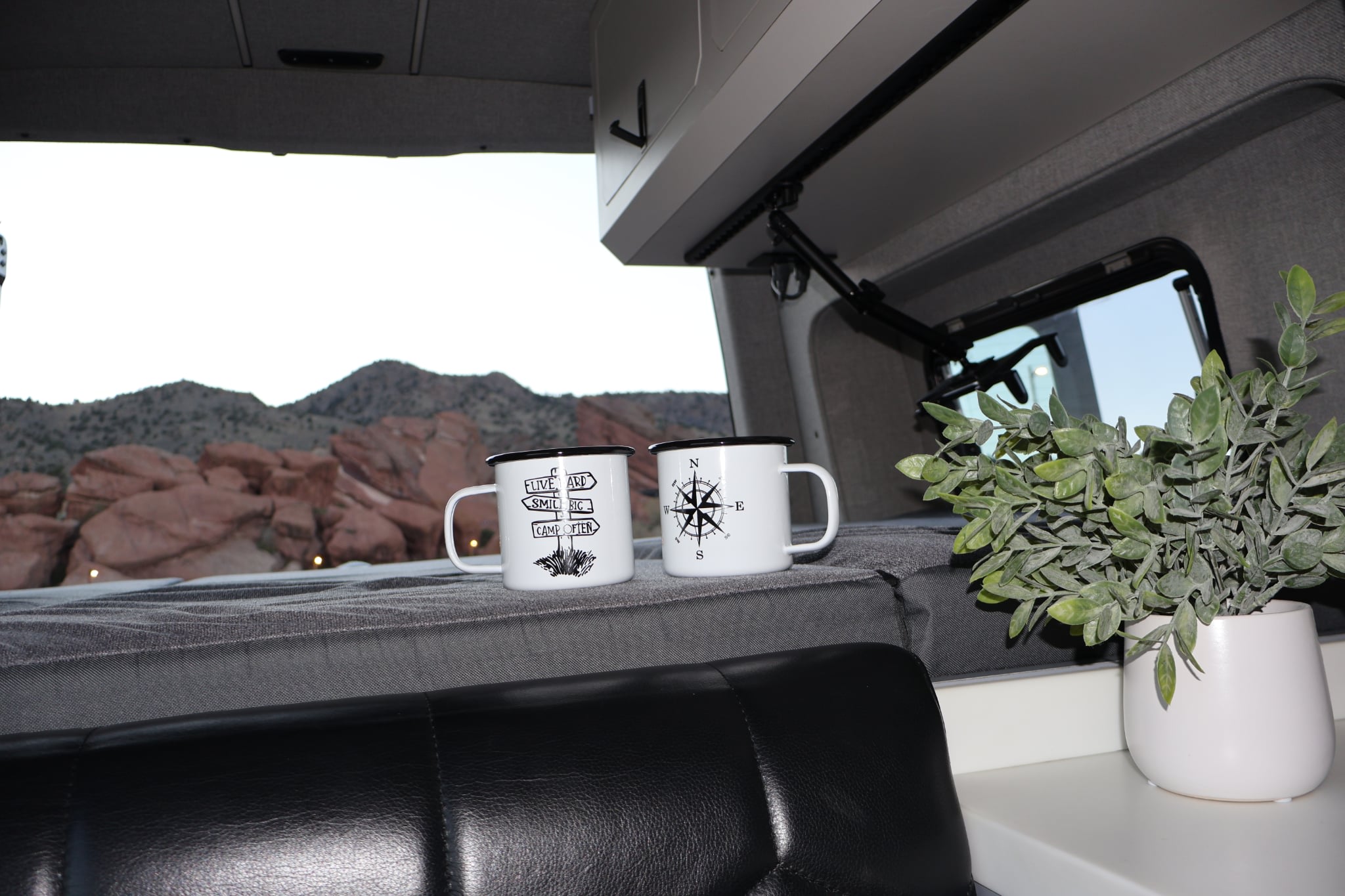 Inside a comfy RV rental, two camping mugs and a plant sit on a bed. Scenic mountains and red rocks are visible through the window.. Remote Vans Oasis 2021