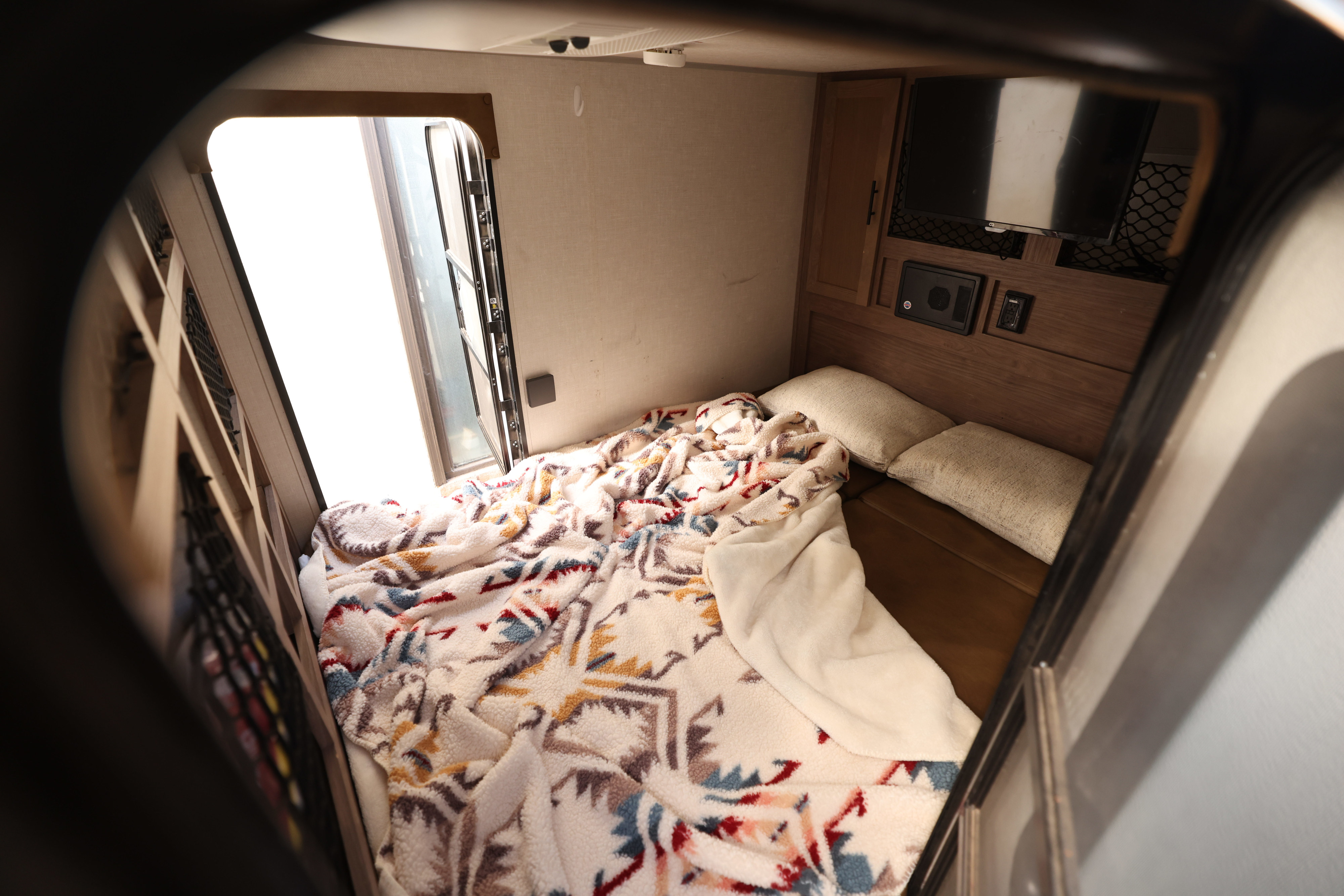 Inside a cozy RV rental bedroom: bed with a colorful blanket, pillows, bright window, TV, and storage.. No Boundaries 10 2020
