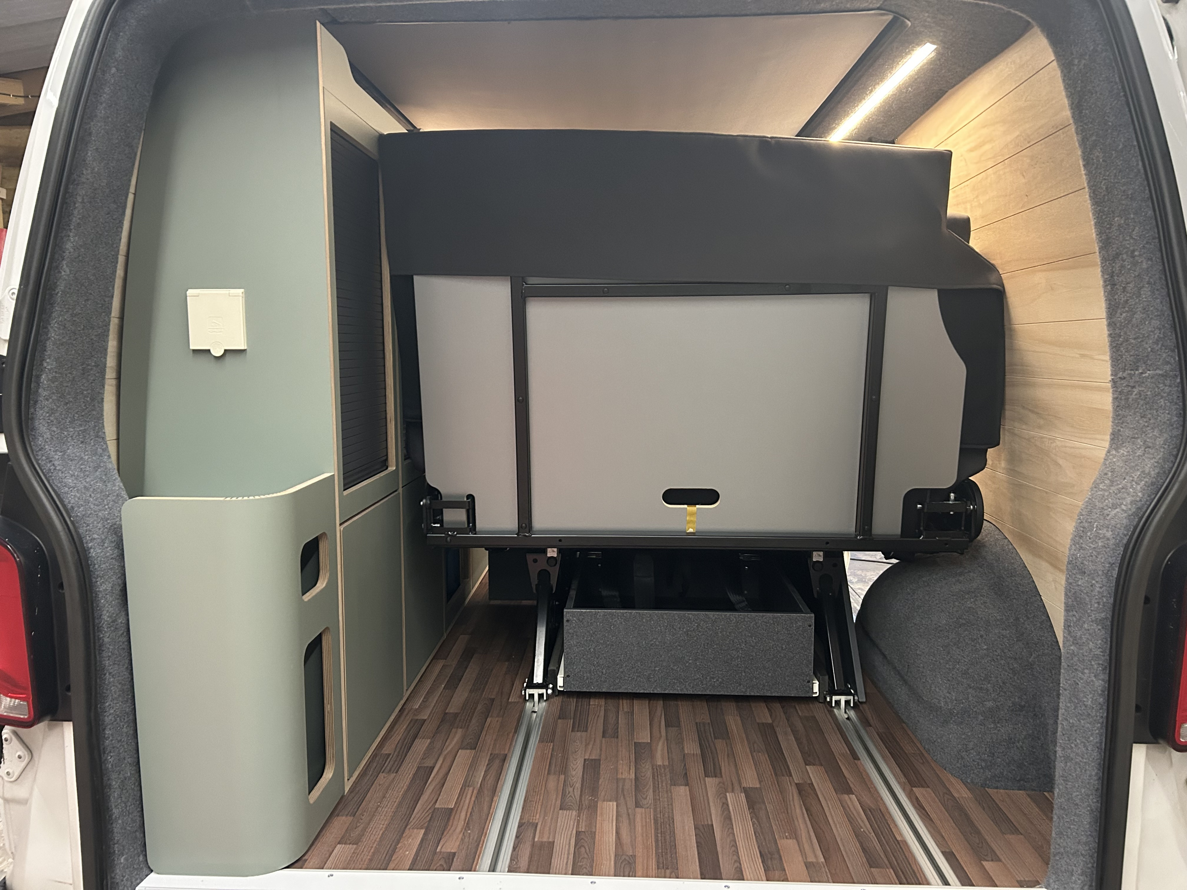 RV rental camper van's rear interior: a compact space with a folded bed, pull-out storage, sage green cabinetry, and wooden walls.. Volkswagen T6 transporter 2021