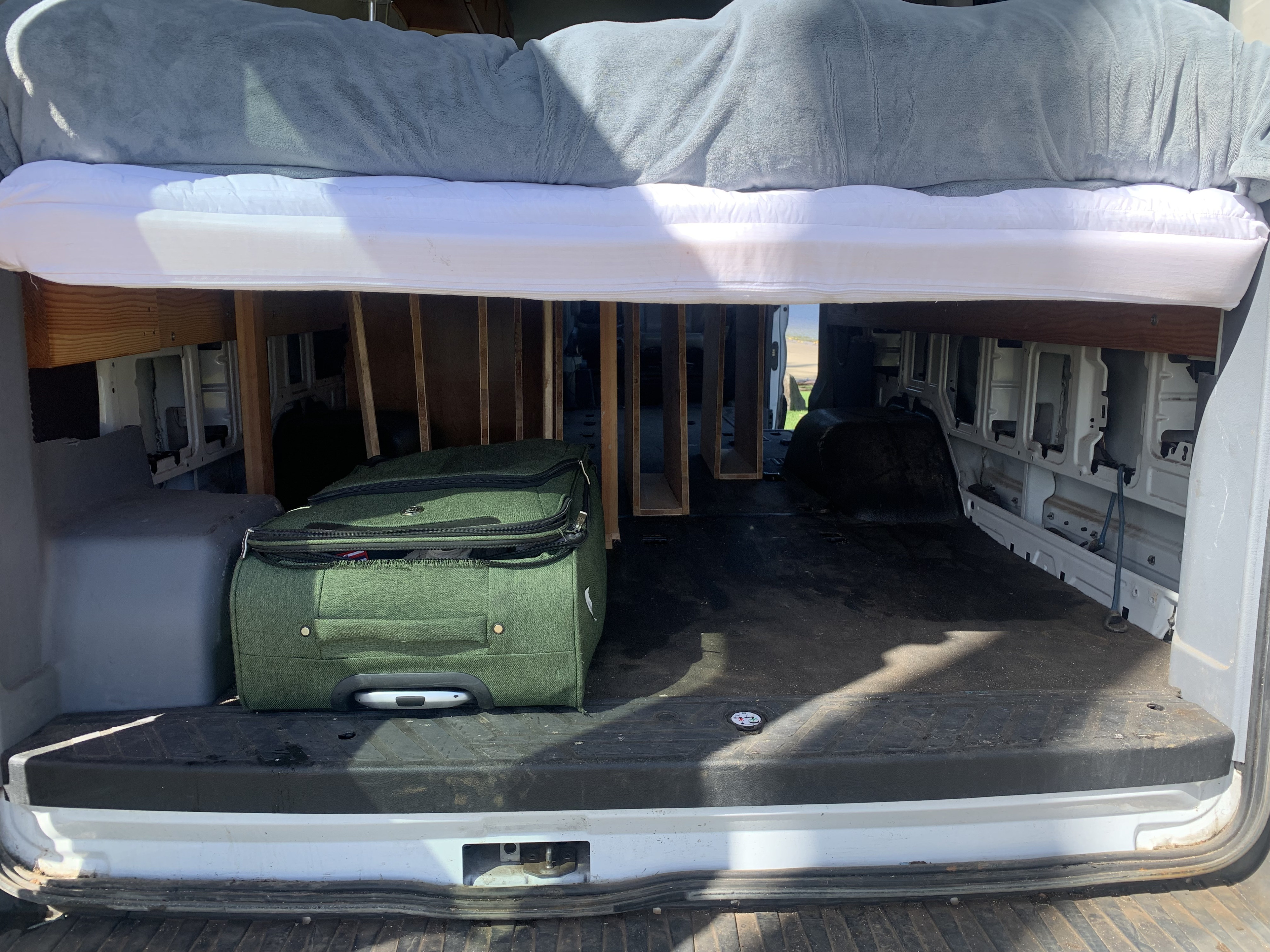 Inside an RV rental van showing a raised bed with mattress, blanket, and ample under-bed storage for luggage like the green suitcase.. ford transit 2016