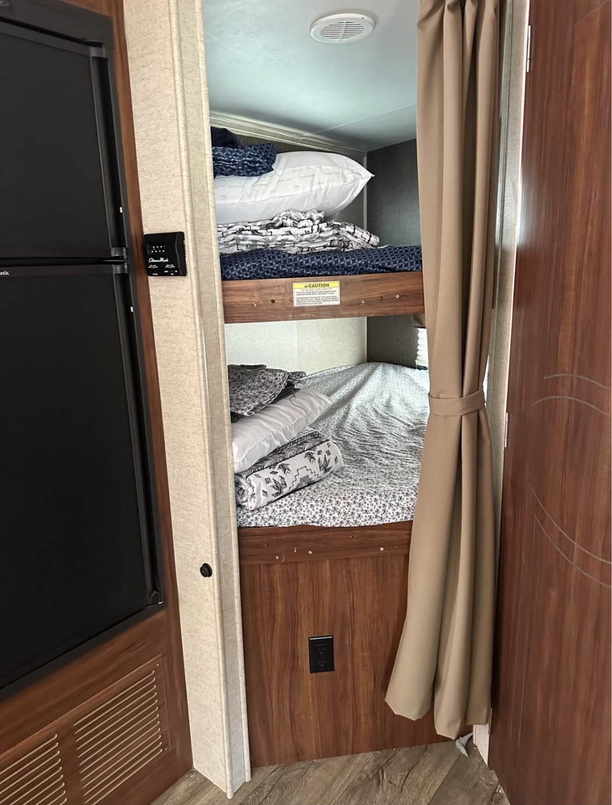 Inside an rv rental: cozy bunk beds with pillows, blankets & curtain. A fridge & controls are to the left.. Heartland RVs Wilderness 2017