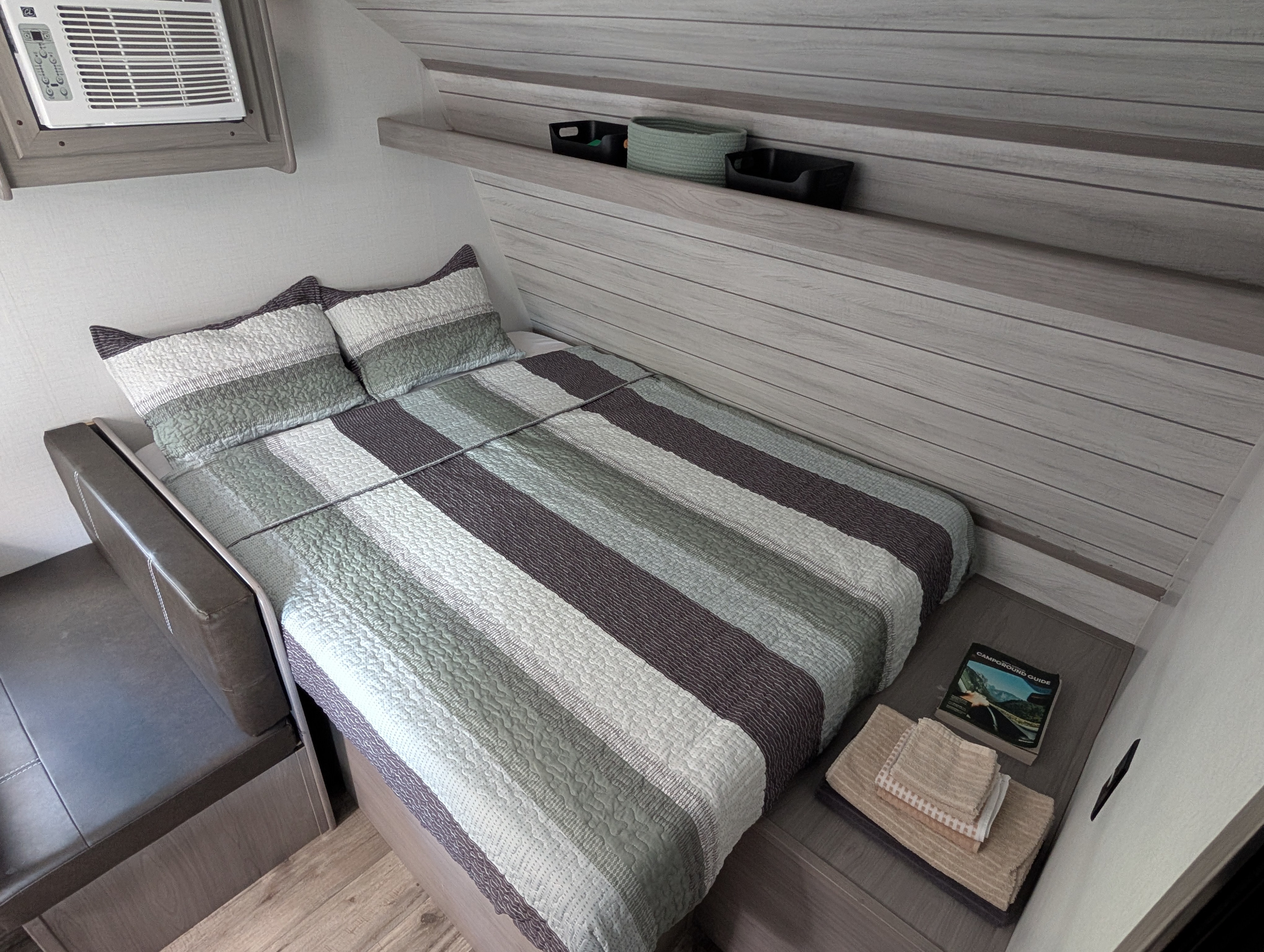 A compact RV rental bedroom featuring a striped bed, mounted AC unit, storage shelf with bins, and a nightstand with a guide book.. Dutchmen Coleman Lantern LT 2023