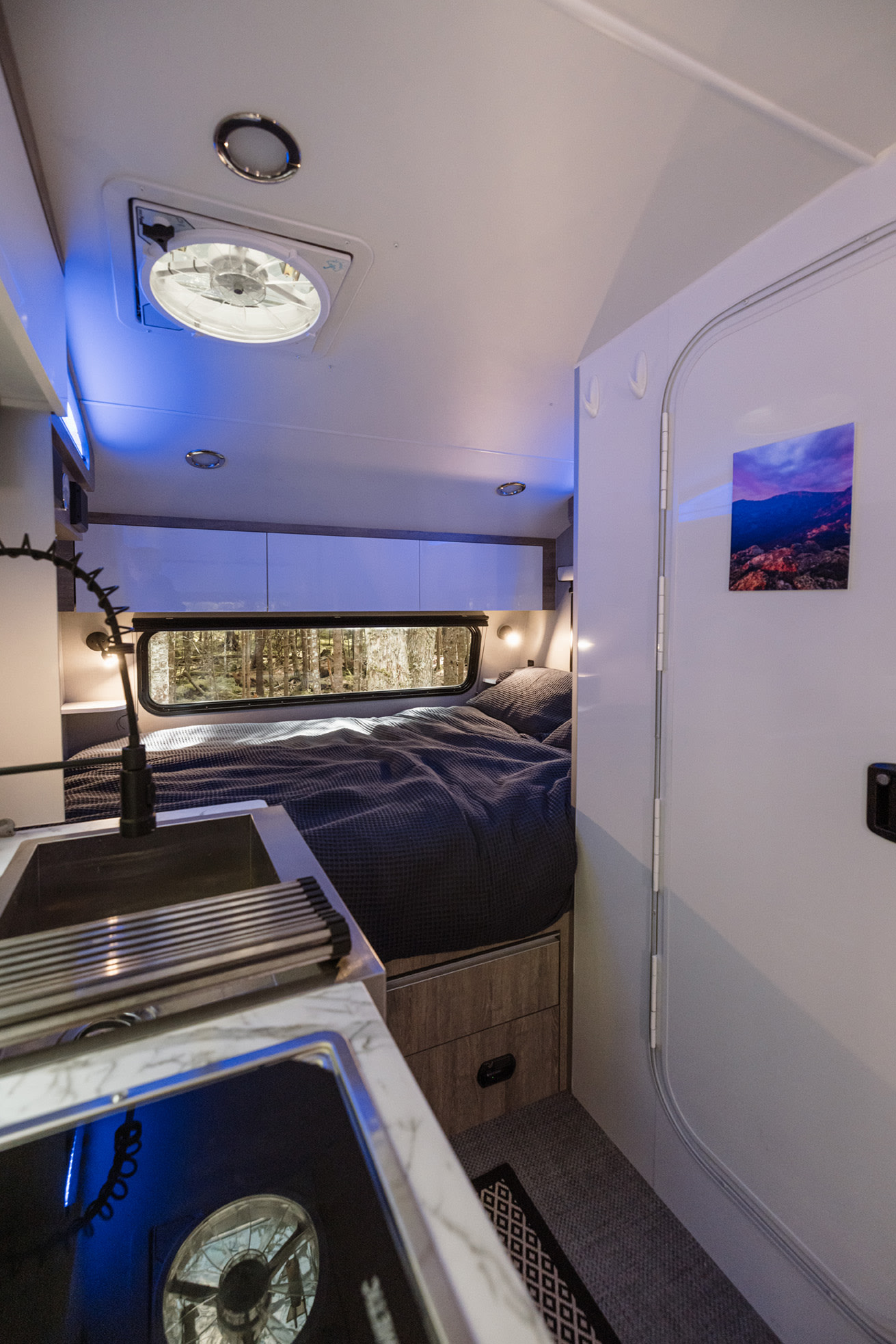 RV rental interior with a cozy bed, compact kitchen, blue accent lighting, and forest view.. InTech Sol Horizon 2025