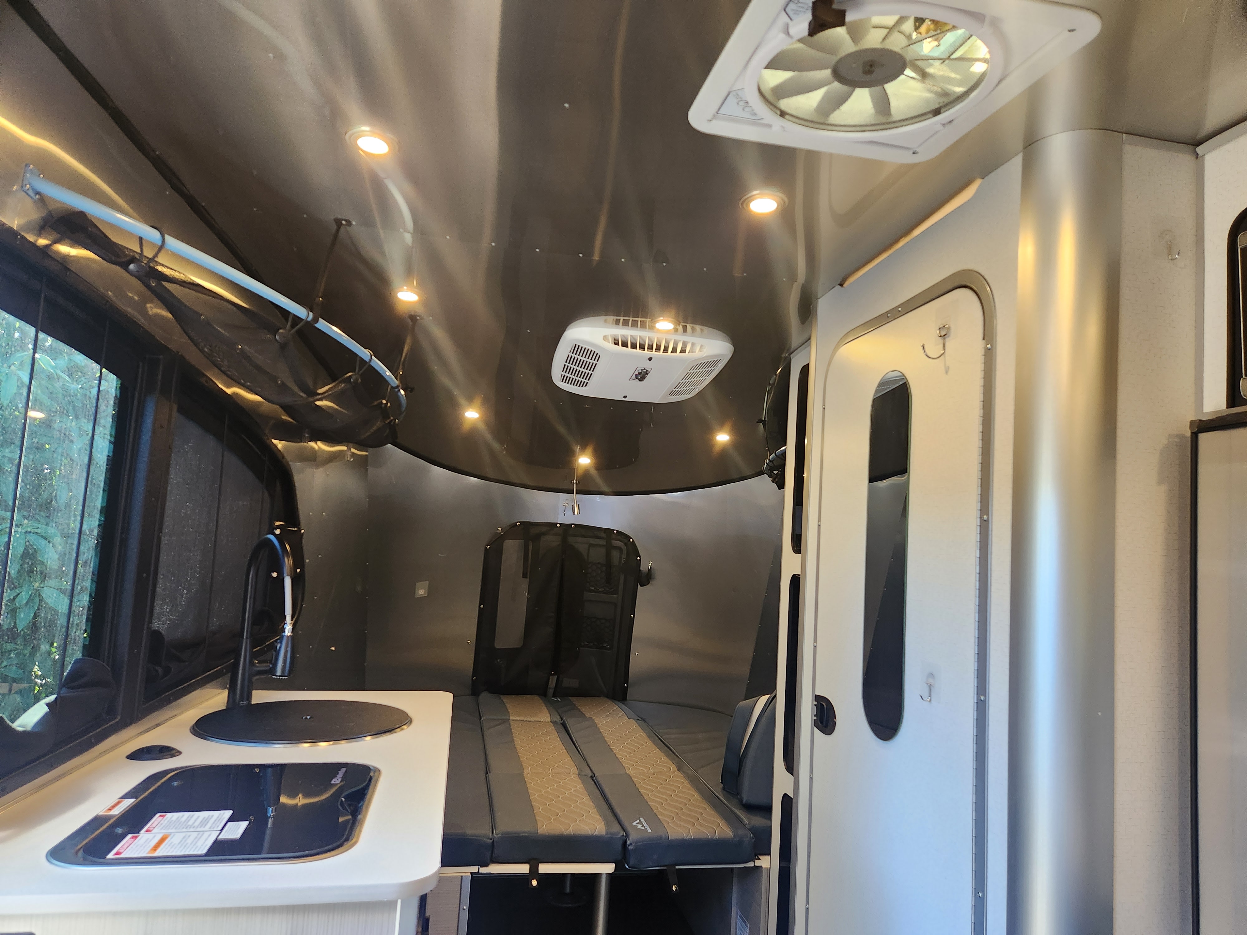 Bright RV rental interior showing kitchenette with sink, fold-down bed, overhead lights, AC, and ceiling fan.. Airstream Basecamp 2025