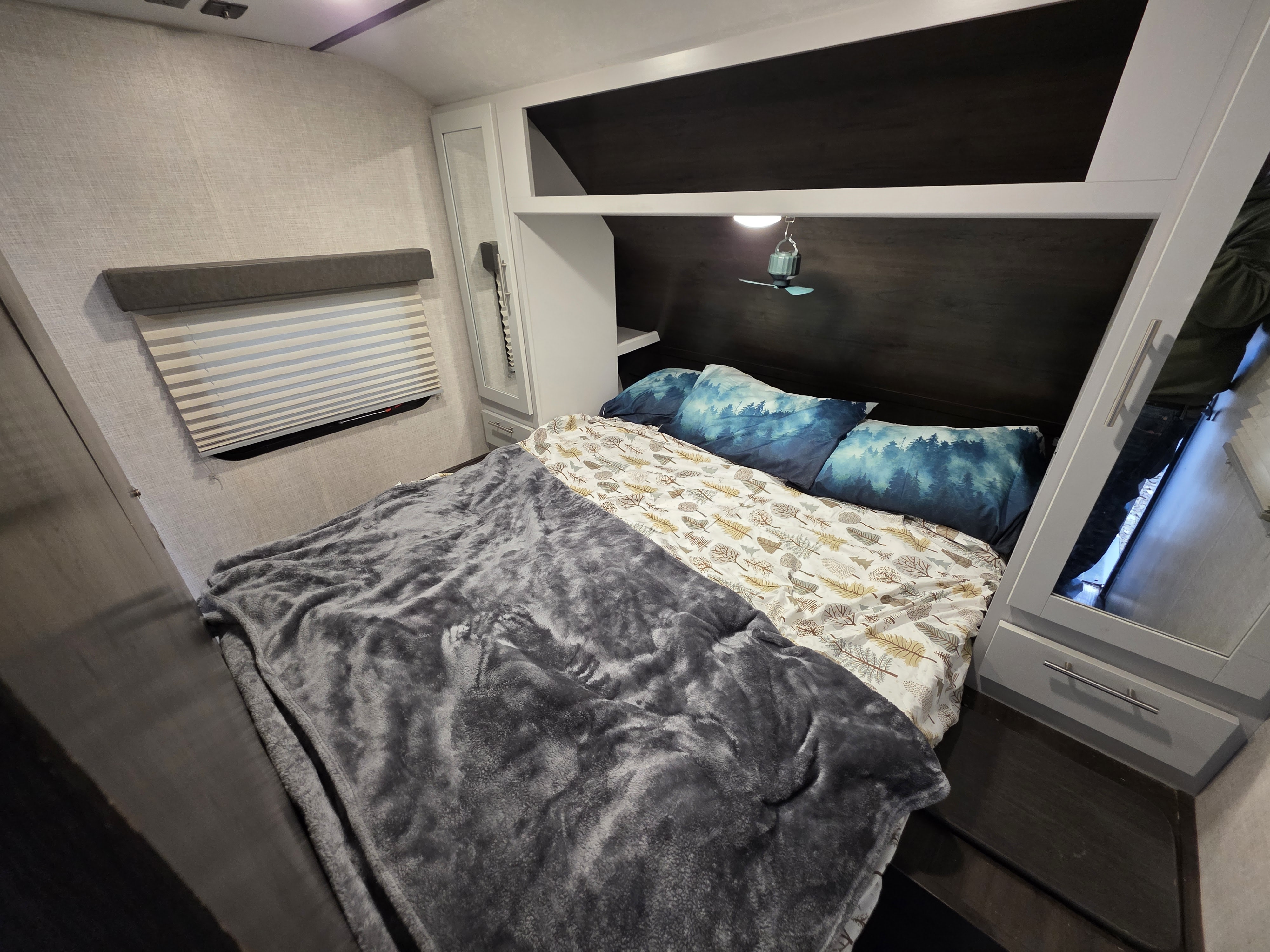 Cozy RV rental bedroom with a bed featuring gray blanket, blue forest pillows, window blinds, and functional overhead storage.. Keystone RV Bullet Ultra Lite 2020