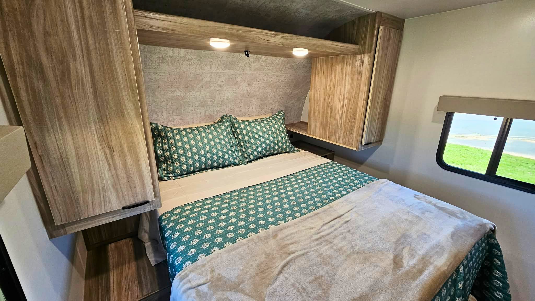 Cozy RV rental bedroom featuring patterned bedding, overhead storage, and a scenic window view of green landscape and water.. Winnebago Access 2024