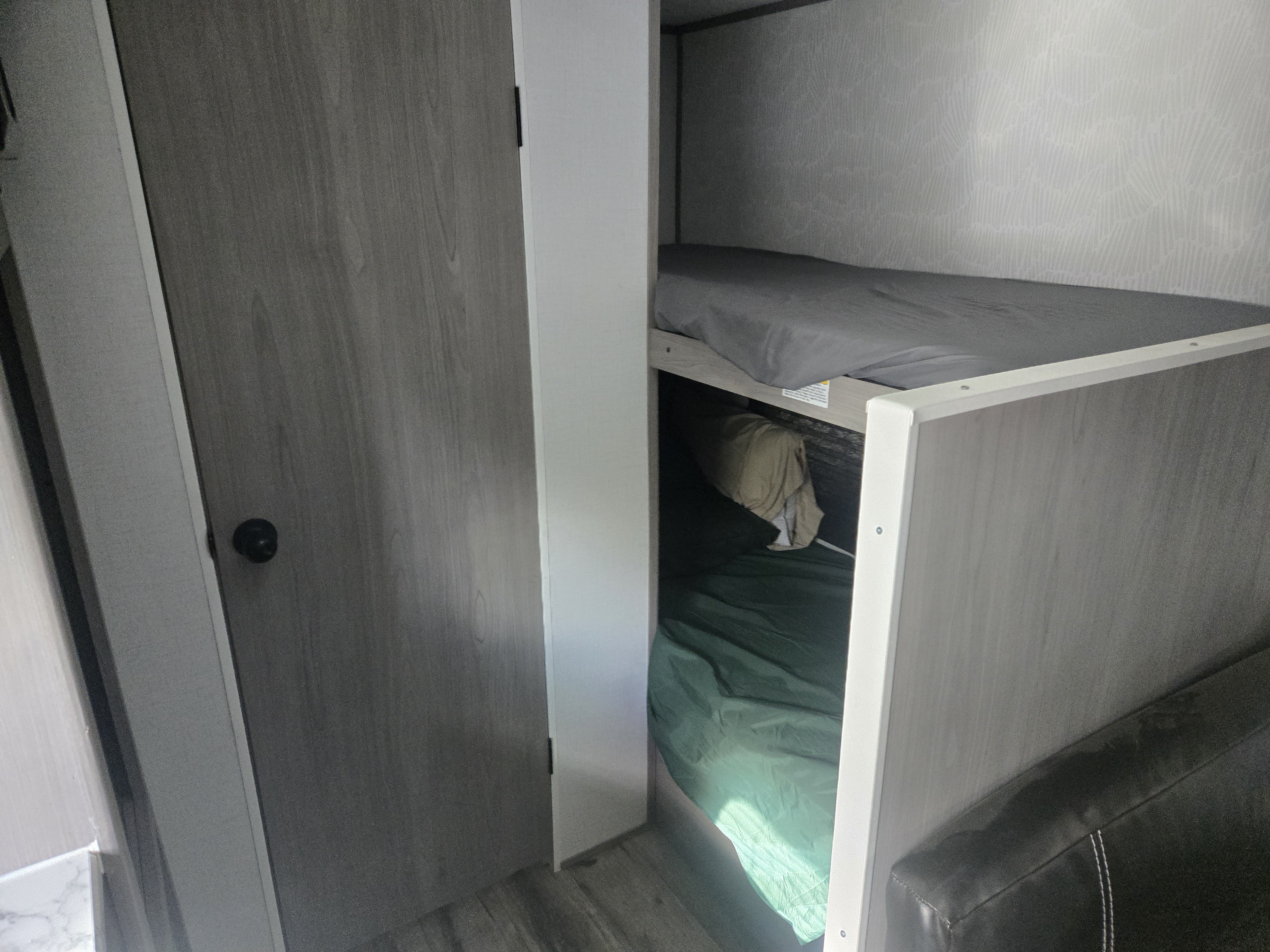 An RV rental interior with bunk beds. The top bed has gray sheets, the bottom has green bedding & a beige item. A door is to the left.. Dutchmen Cmb1724 2024