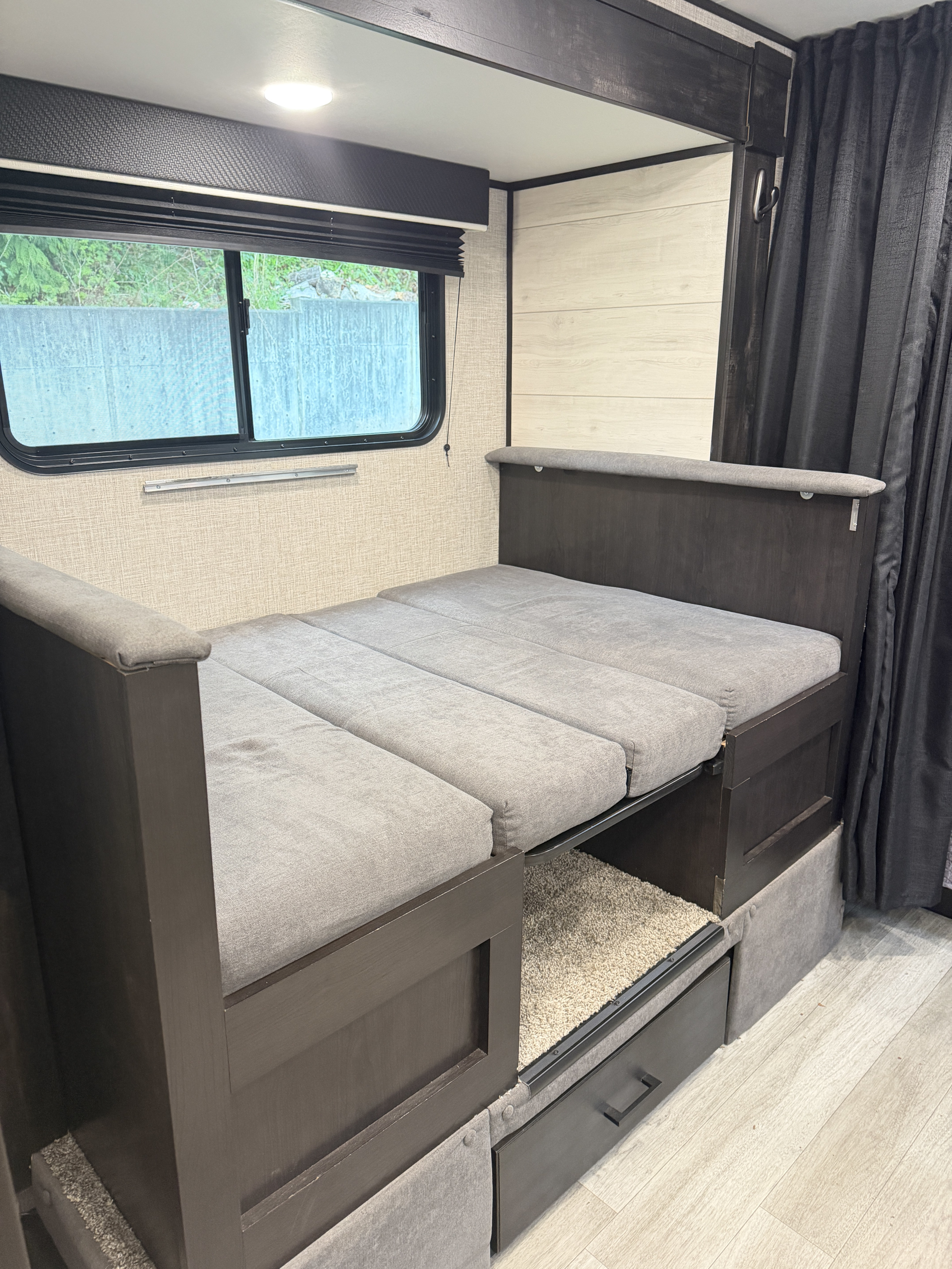 RV rental interior features a convertible gray cushion bed, dark wood accents, a window, and storage drawers.. Jayco Jay Flight SLX Baja Edition 2023