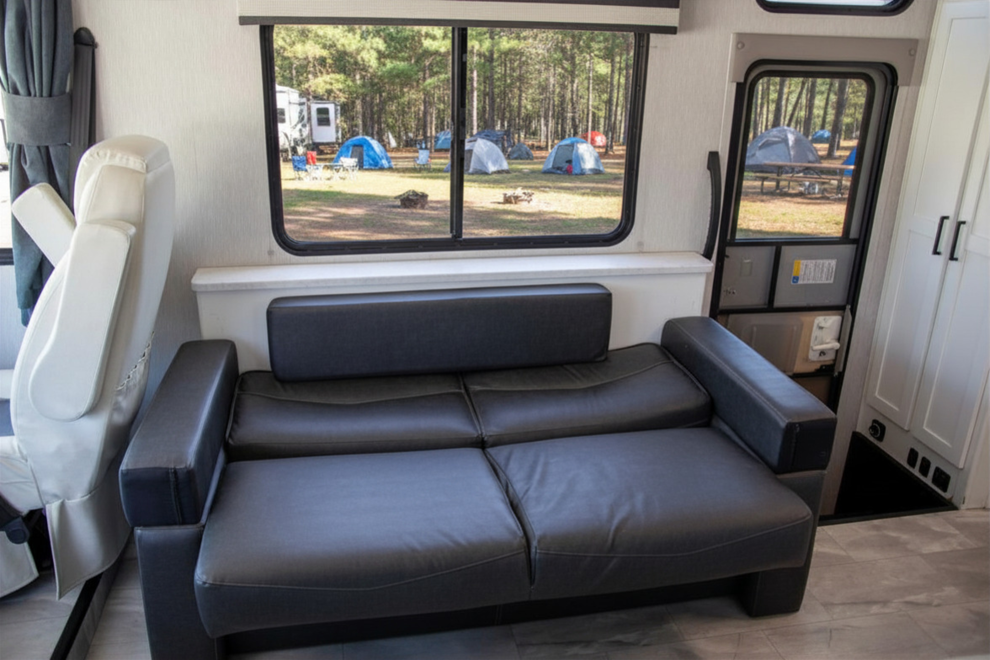 Cozy RV rental interior with a dark sofa. A large window and door reveal a vibrant campground scene with multiple tents among pine trees.. Forest River Coachmen Pursuit 2024