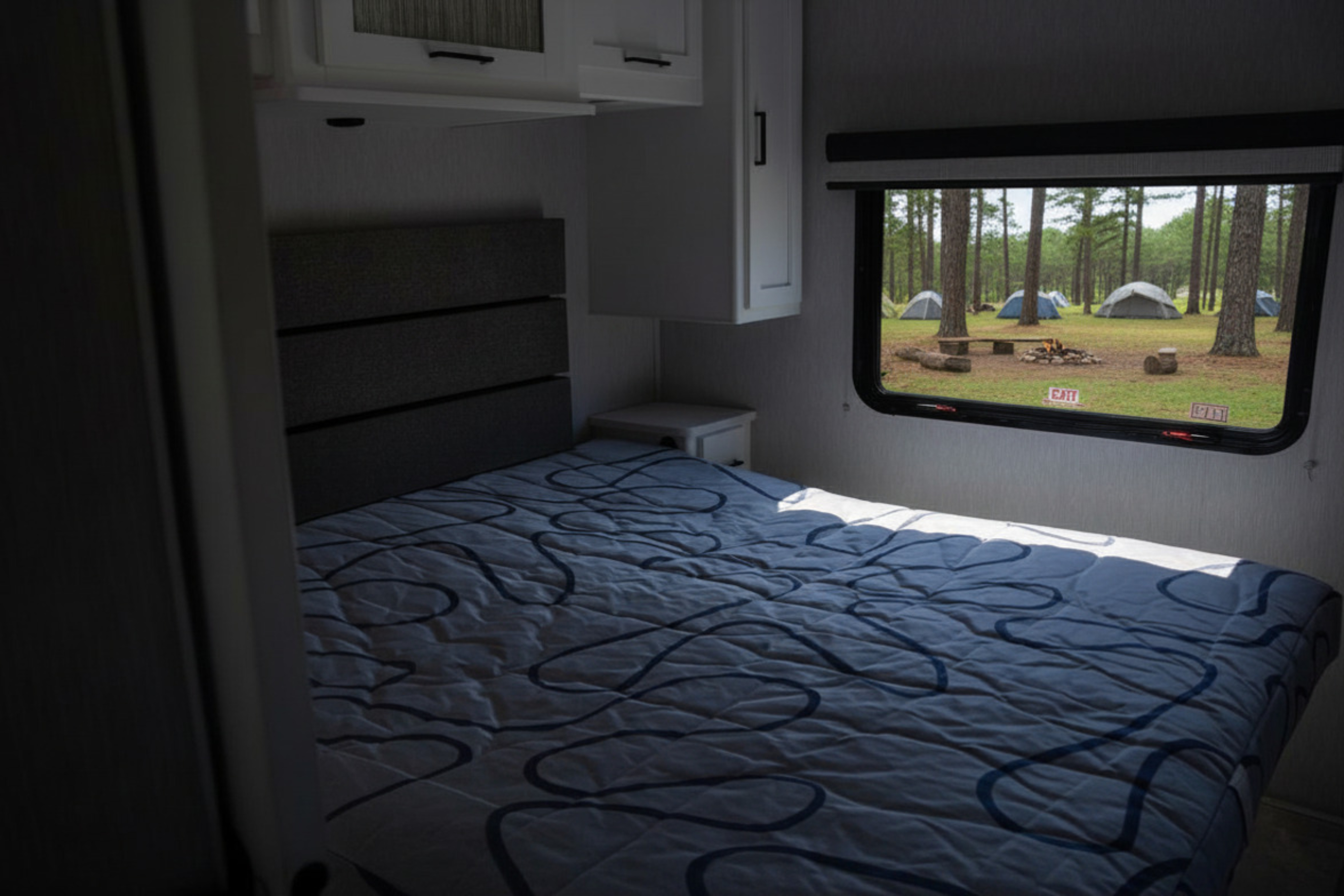 RV rental bedroom with a cozy bed and a window view of a peaceful forest campsite, featuring tents, trees, and a campfire.. Forest River Coachmen Pursuit 2024