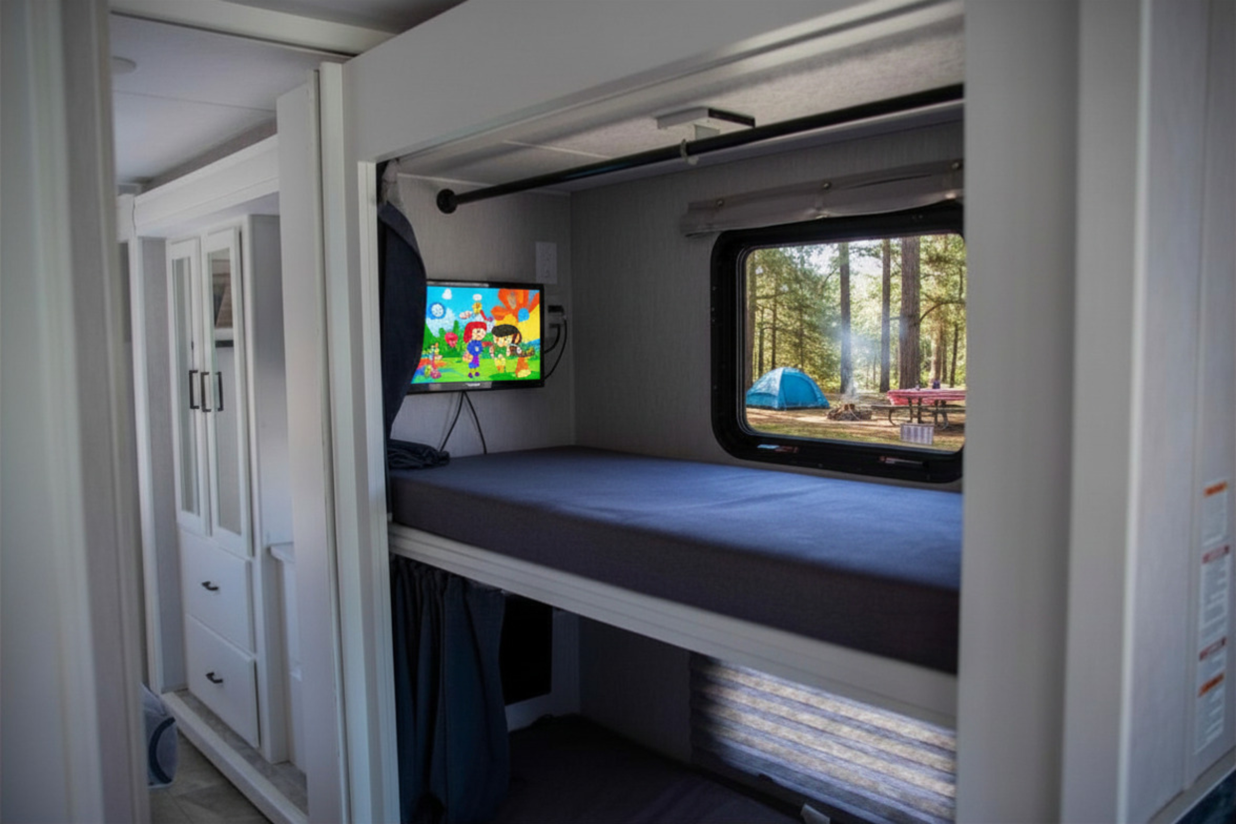 An RV rental's bunk room with a TV, comfy beds, and a scenic window overlooking a campsite with a blue tent.. Forest River Coachmen Pursuit 2024