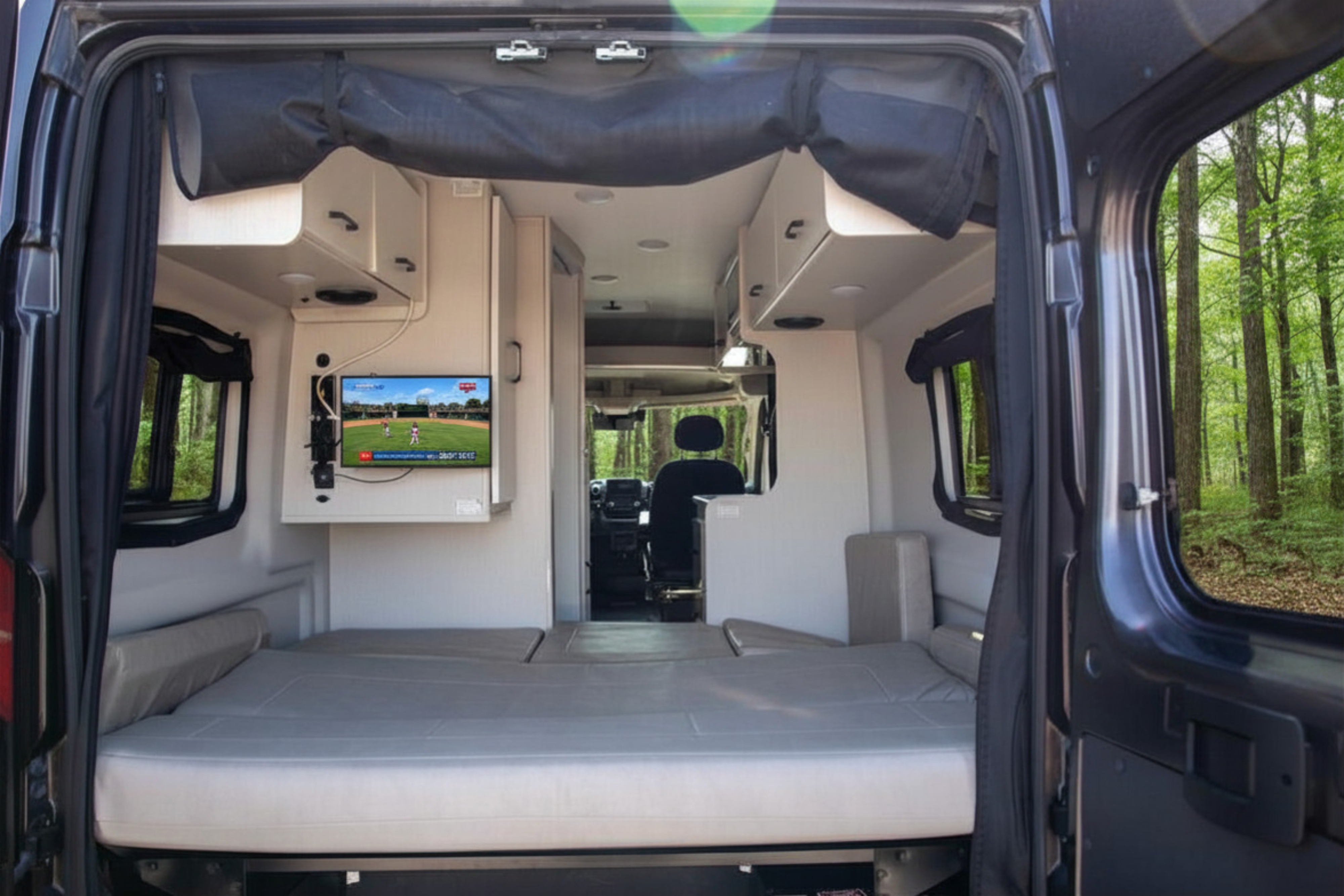 Inside an RV rental: a cozy camper van with a bed, TV playing baseball, and a beautiful forest view outside the open rear doors.. Thor Twist 2023
