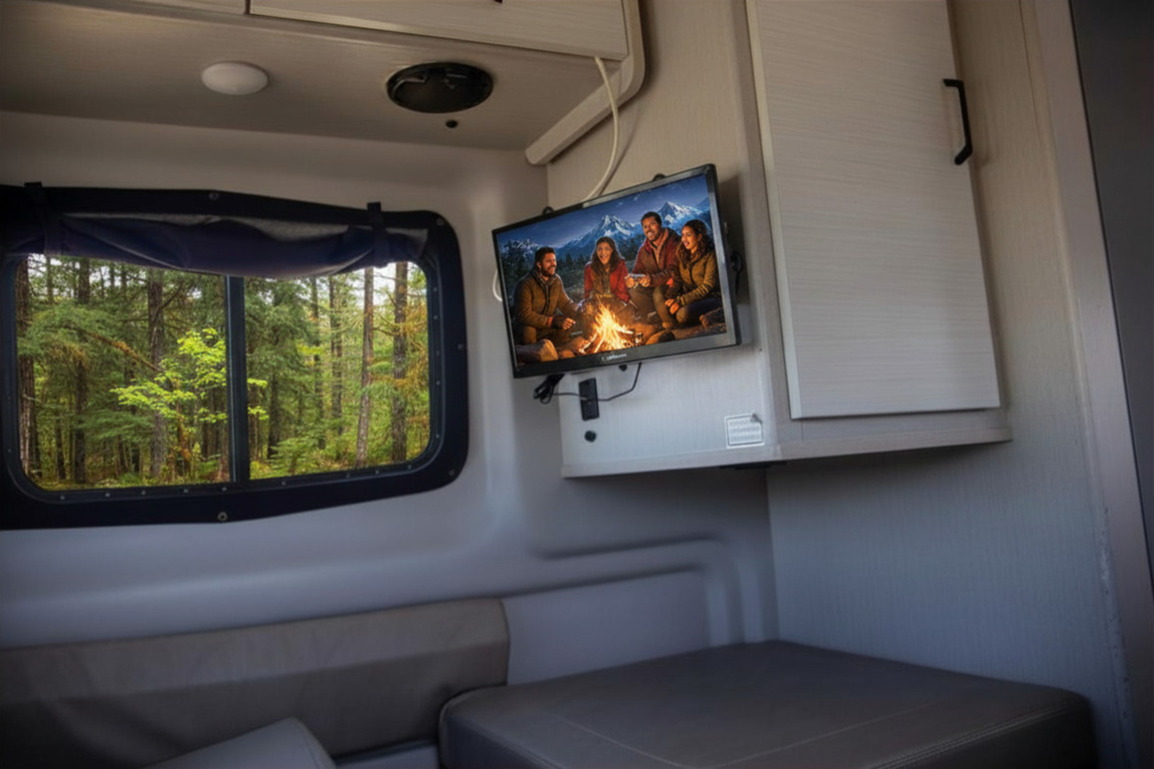 Inside an RV rental: TV shows campers by a fire, a window overlooks a serene forest, with comfy seating & overhead storage.. Thor Twist 2023