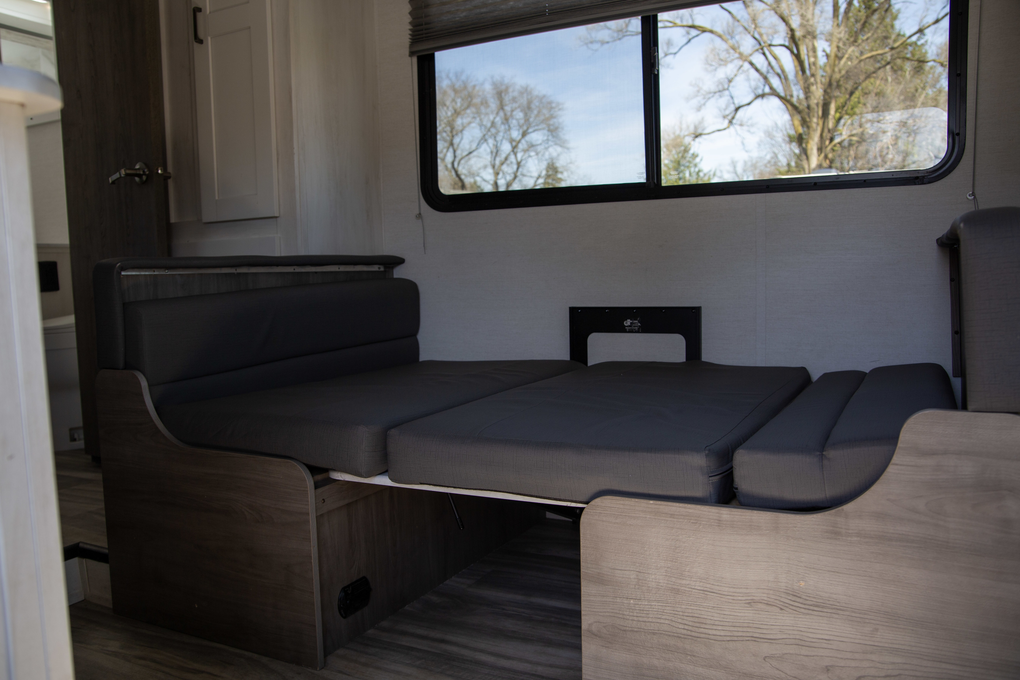 Cozy RV rental interior with dinette converted to a bed, gray cushions, and a large window view of trees.. Gulf Stream Conquest 2024